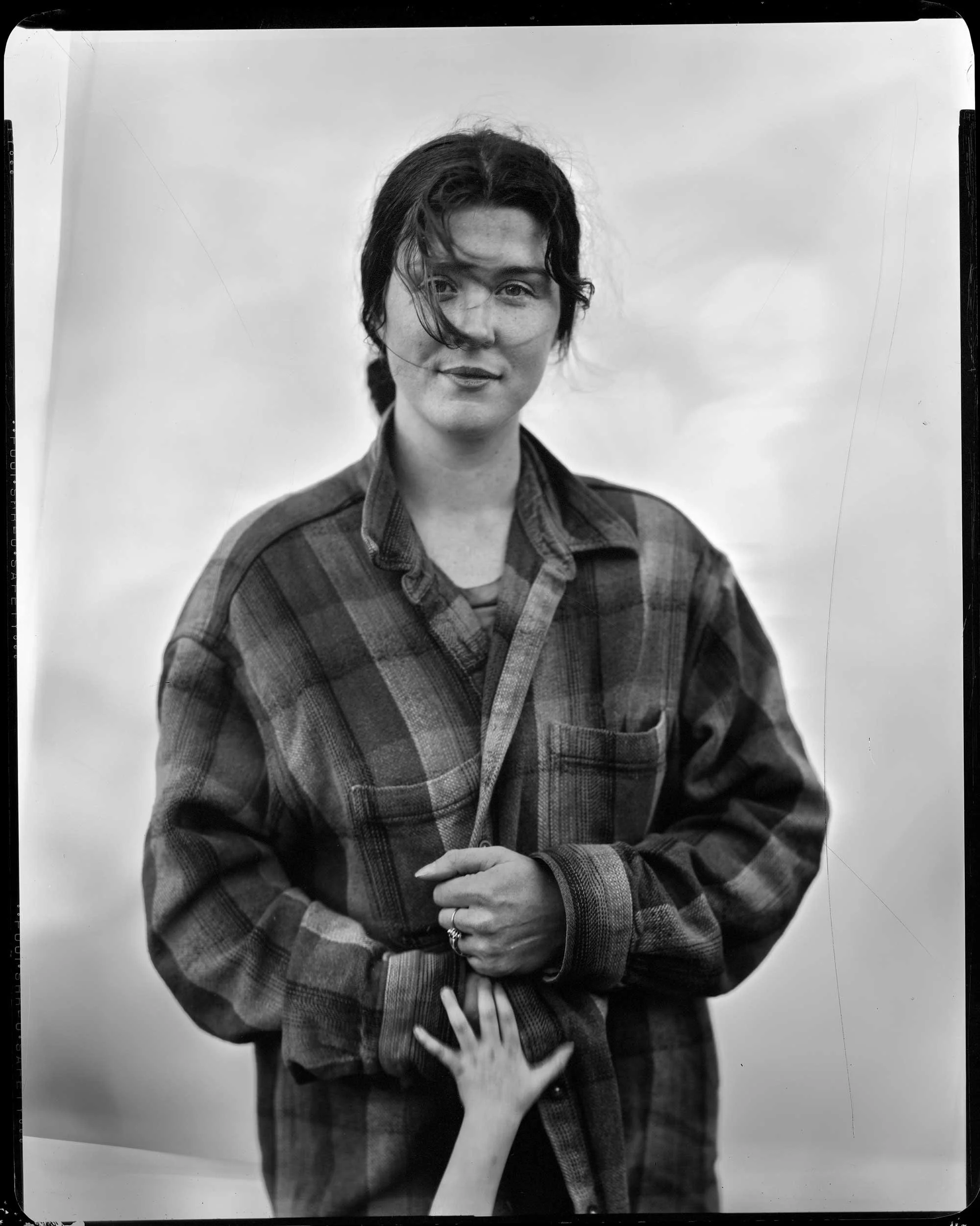A striking black and white studio portrait of a woman wearing an oversized plaid shirt, with a young child's hand reaching up into the frame to touch her hand, set against a soft sky backdrop in Appalachia.