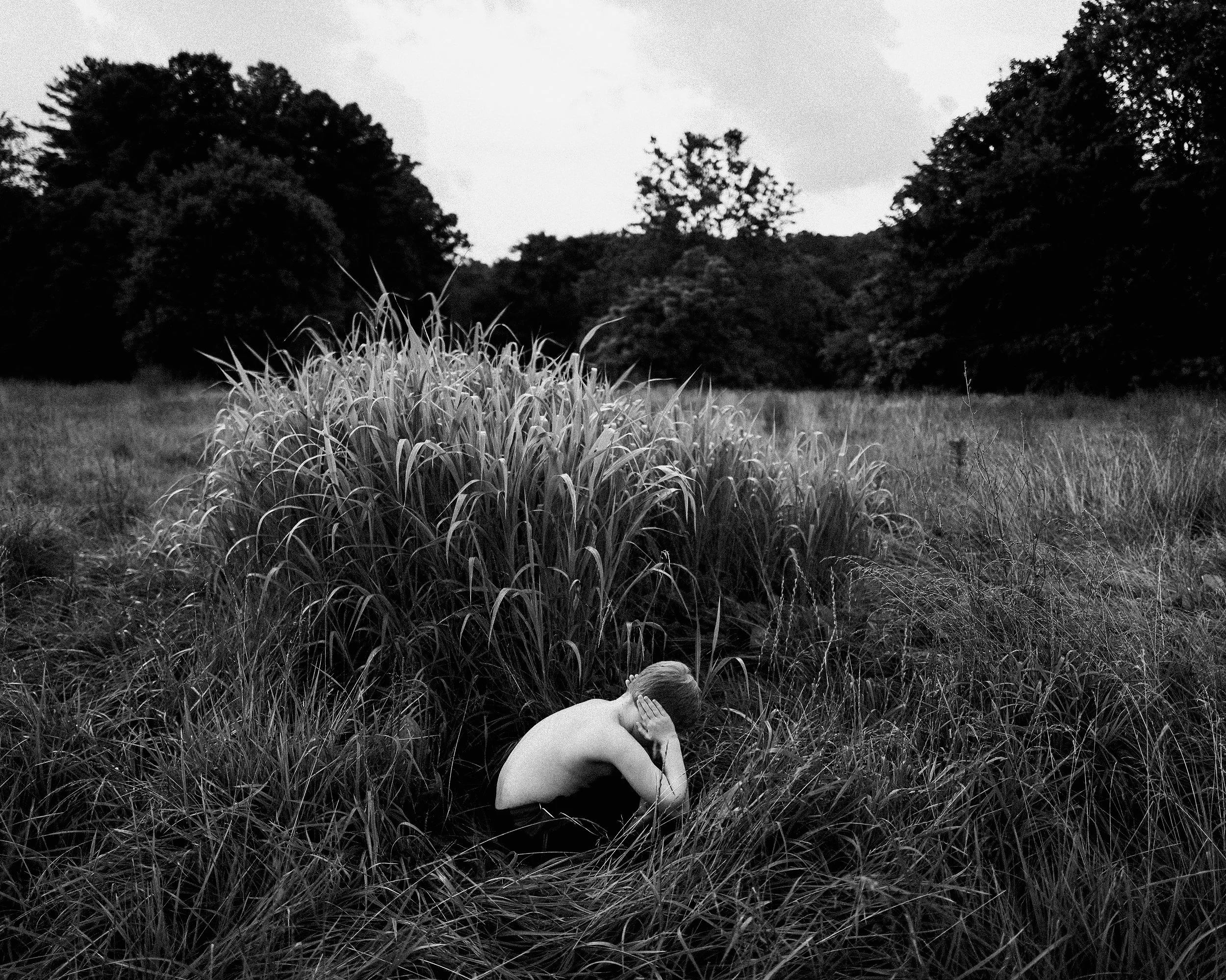 A poignant black and white fine art photograph of a young, shirtless boy crouched down inside a thick patch of tall, wild grass in an open field, holding his hands over his ears with his head down, set against a background of dense trees.