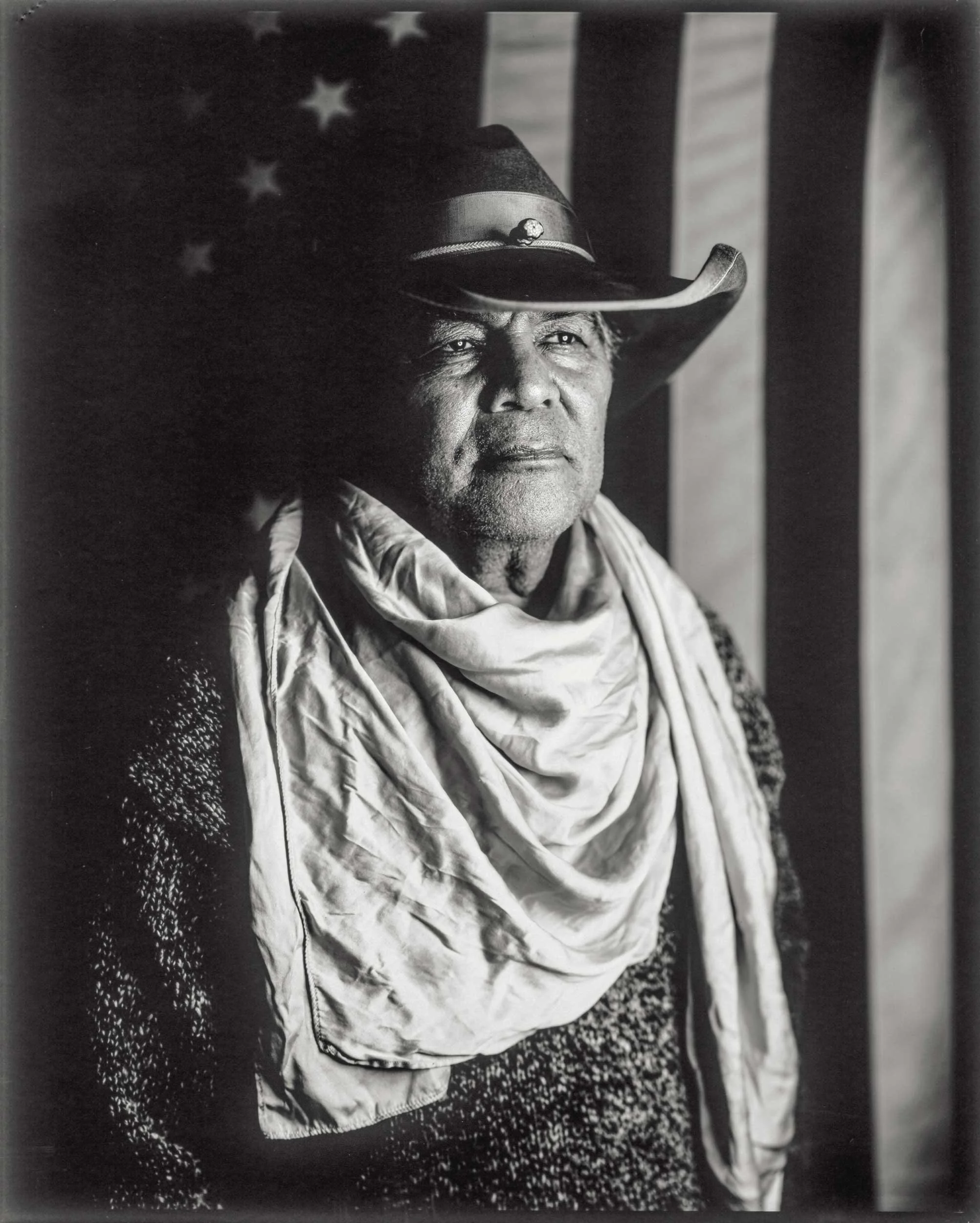 A dramatic black and white studio portrait of a man wearing a cowboy hat and a draped scarf, looking off-camera against a softly focused American flag background, capturing a face of the Appalachian community in Western North Carolina.