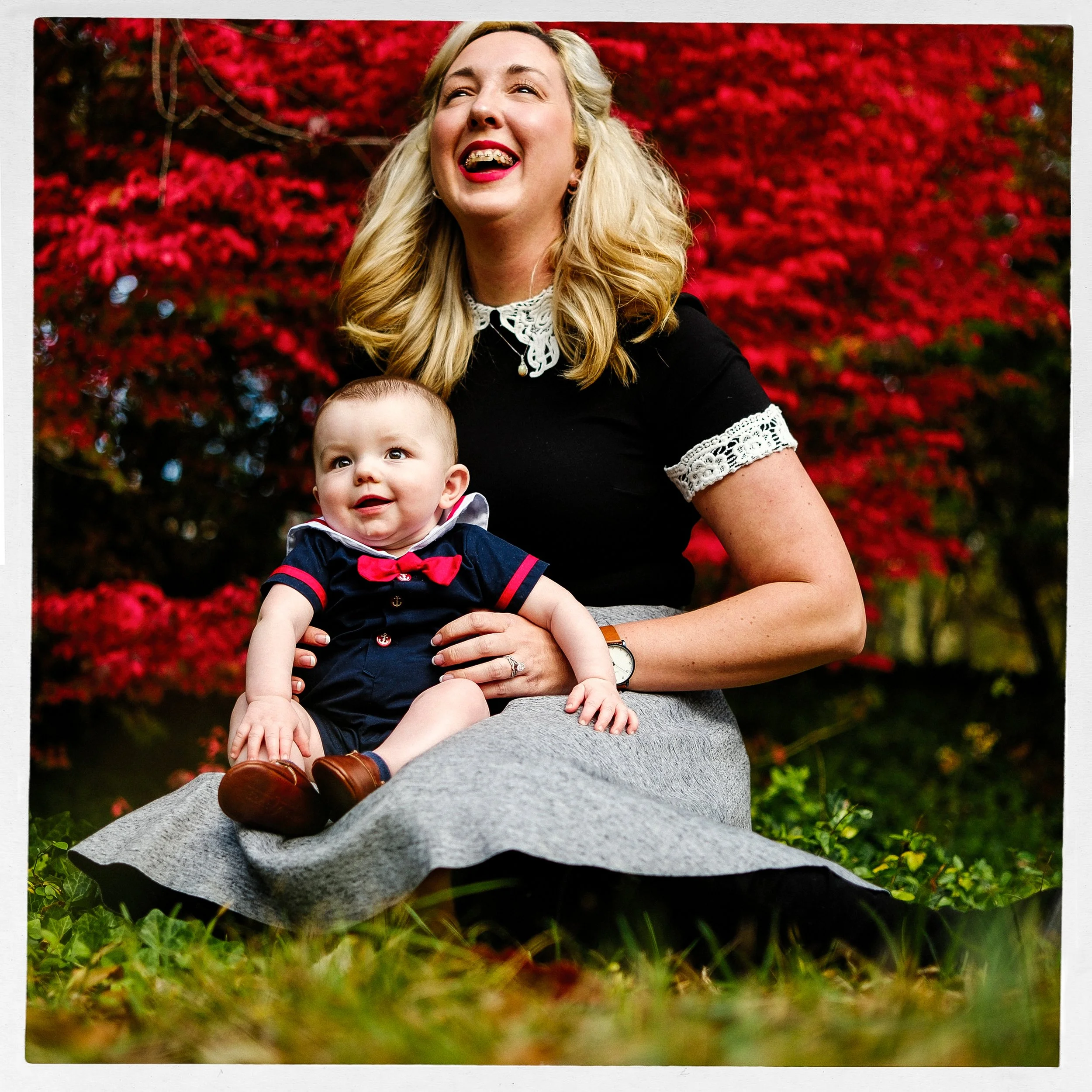 Vibrant, joyful color portrait of a woman laughing happily with a baby sitting on her lap, set against a backdrop of brilliant red autumn leaves in Asheville.