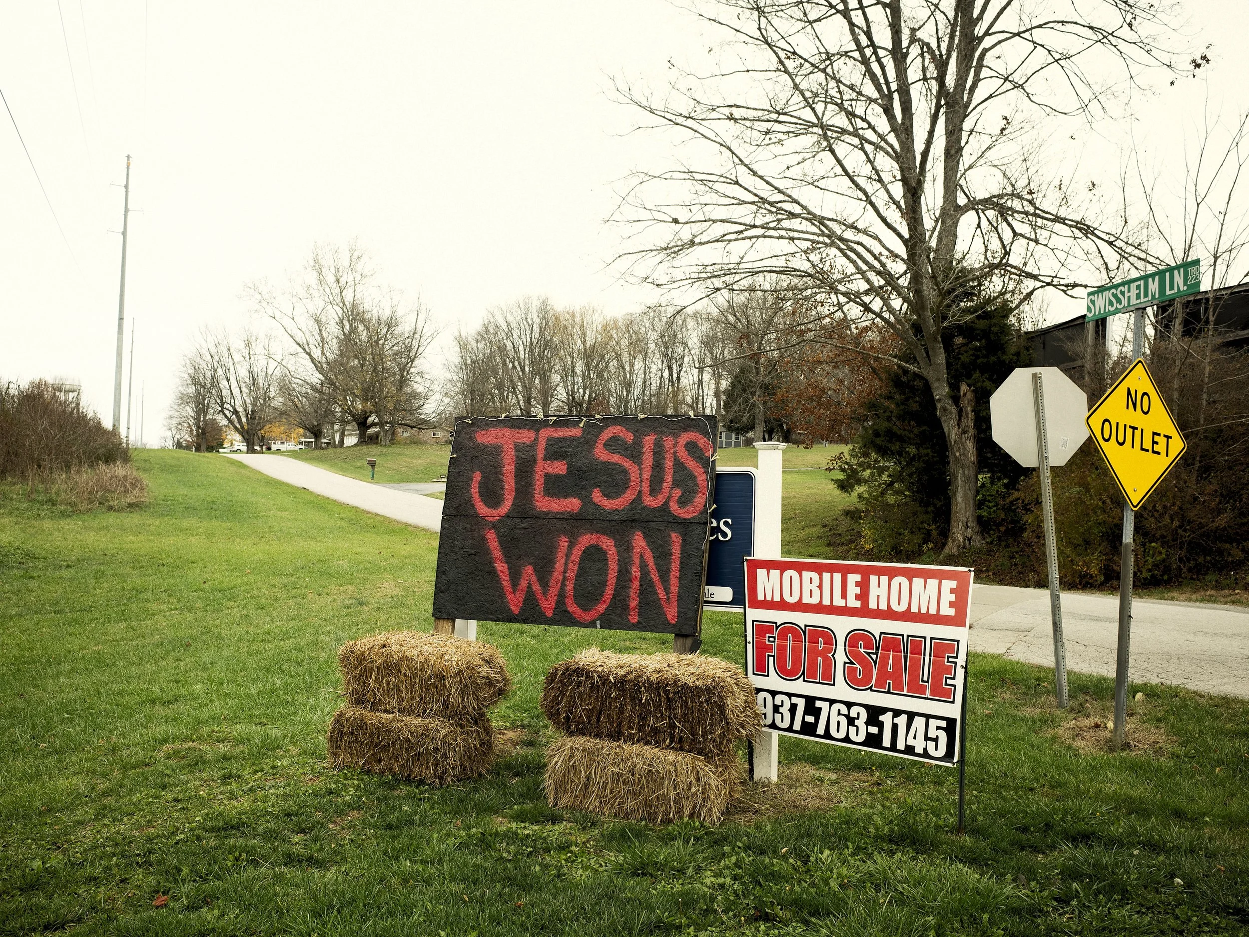 A color environmental photograph of a hand-painted "JESUS WON" sign on hay bales that cover a Harris for President sign, the alongside a "Mobile Home For Sale" sign. The day Trump was elected, from the One Way Out, Lord series by Parker J Pfister