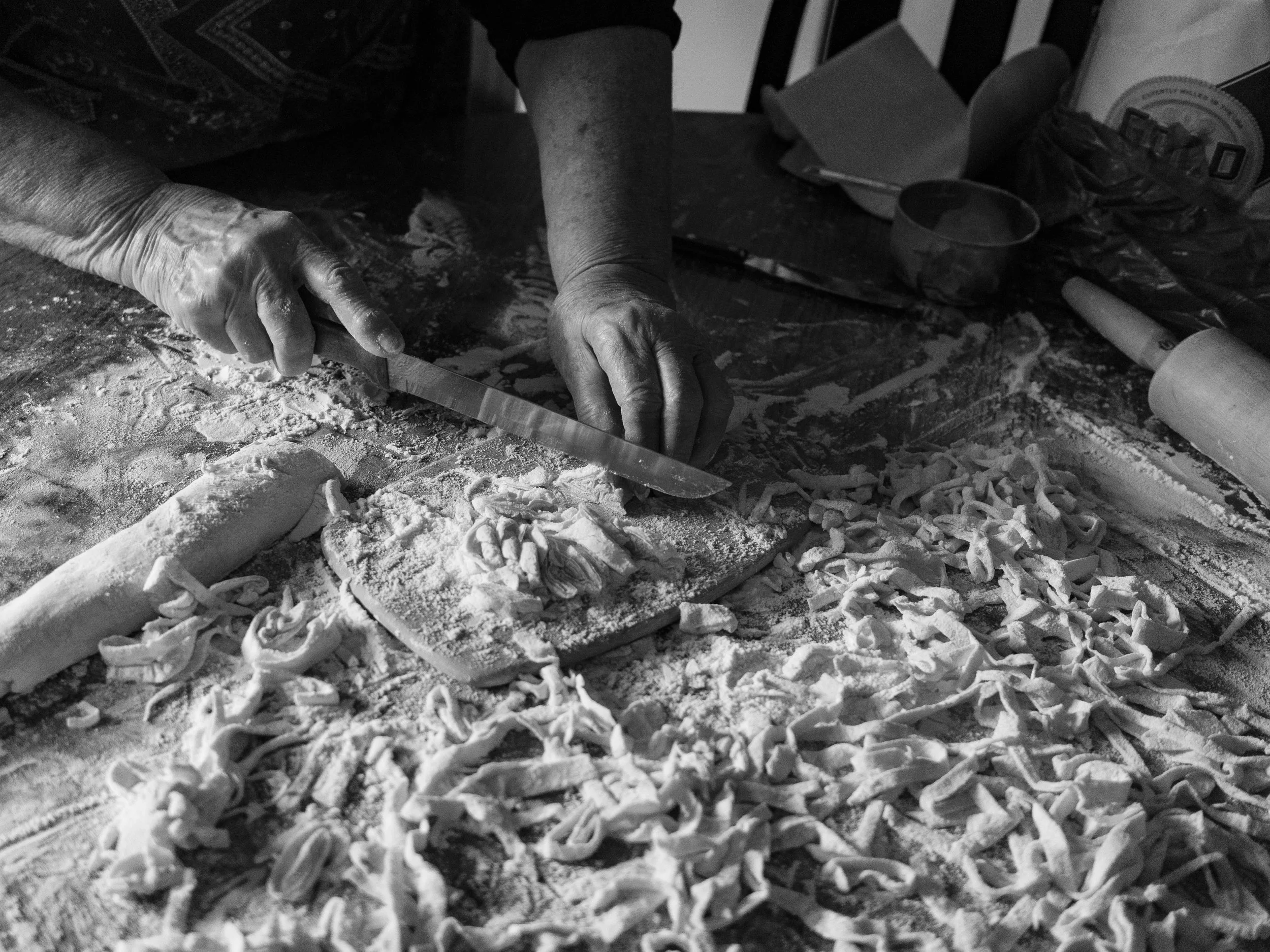 A high-contrast black and white documentary photograph capturing a close-up of a person's hands using a knife to cut fresh, homemade noodles on a heavily floured wooden table, from One Way Out, Lord by Parker J Pfister.