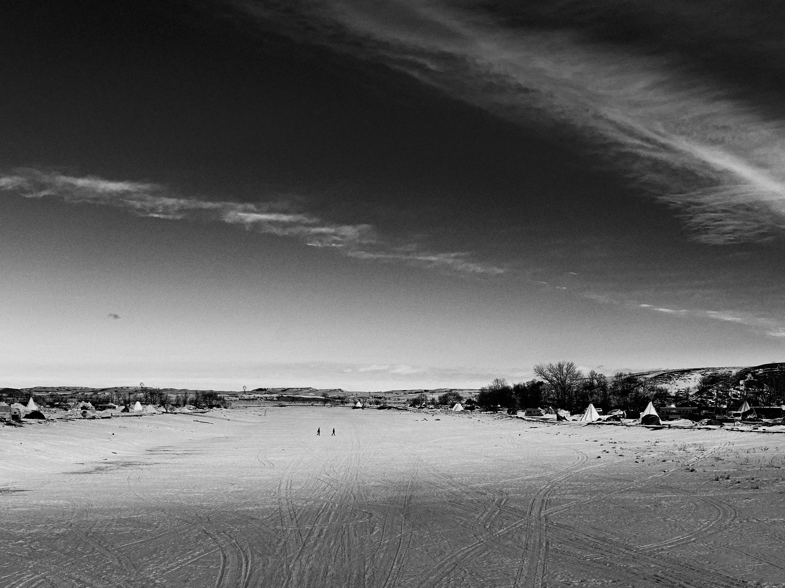 Black and white landscape of a ice-covered Cannonball River at the Standing Rock protest camp, with vast tire tracks in the snow and two distant figures walking under a dramatic winter sky.