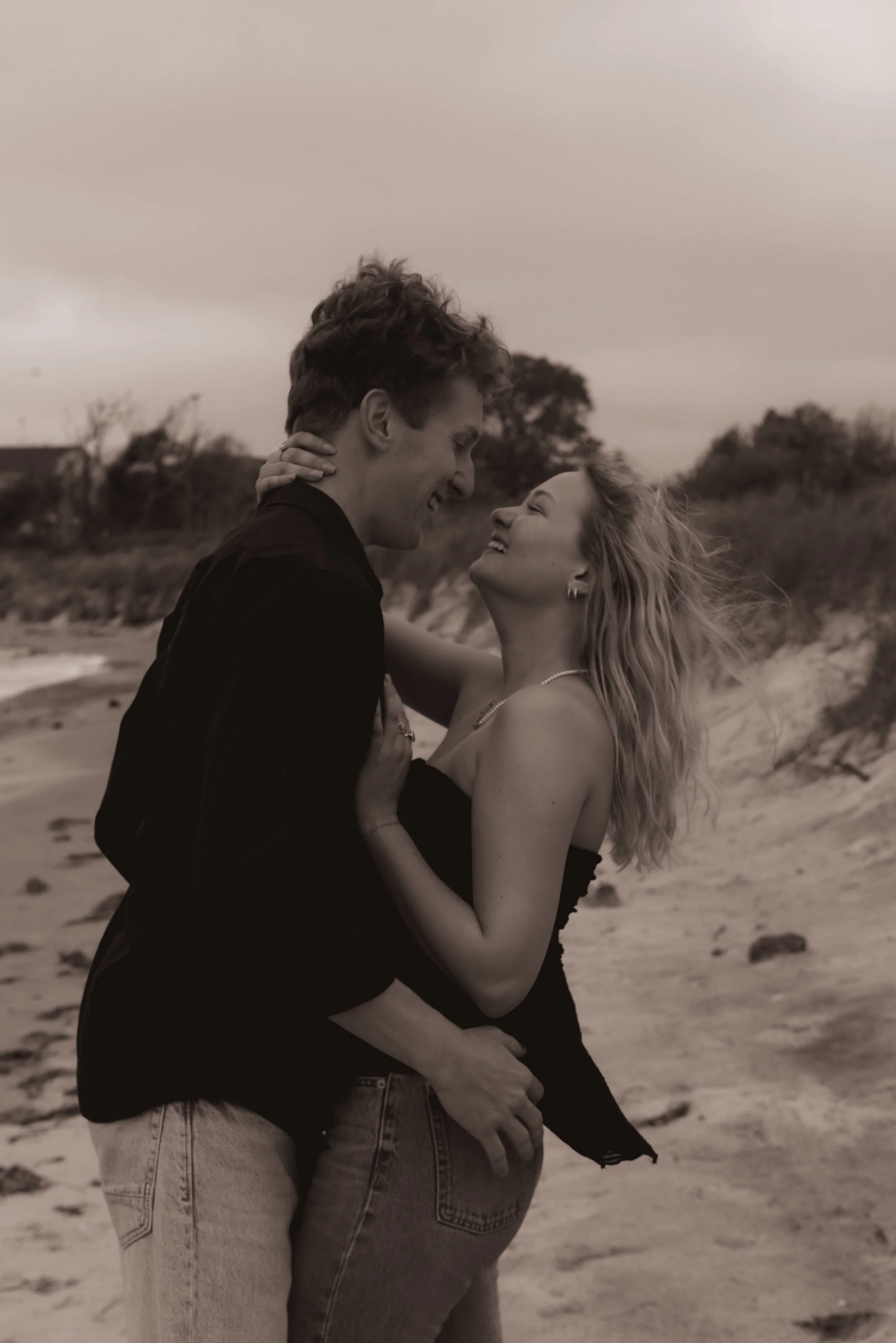 A black and white photo of a couple embracing on a beach, smiling and looking into each other's eyes.