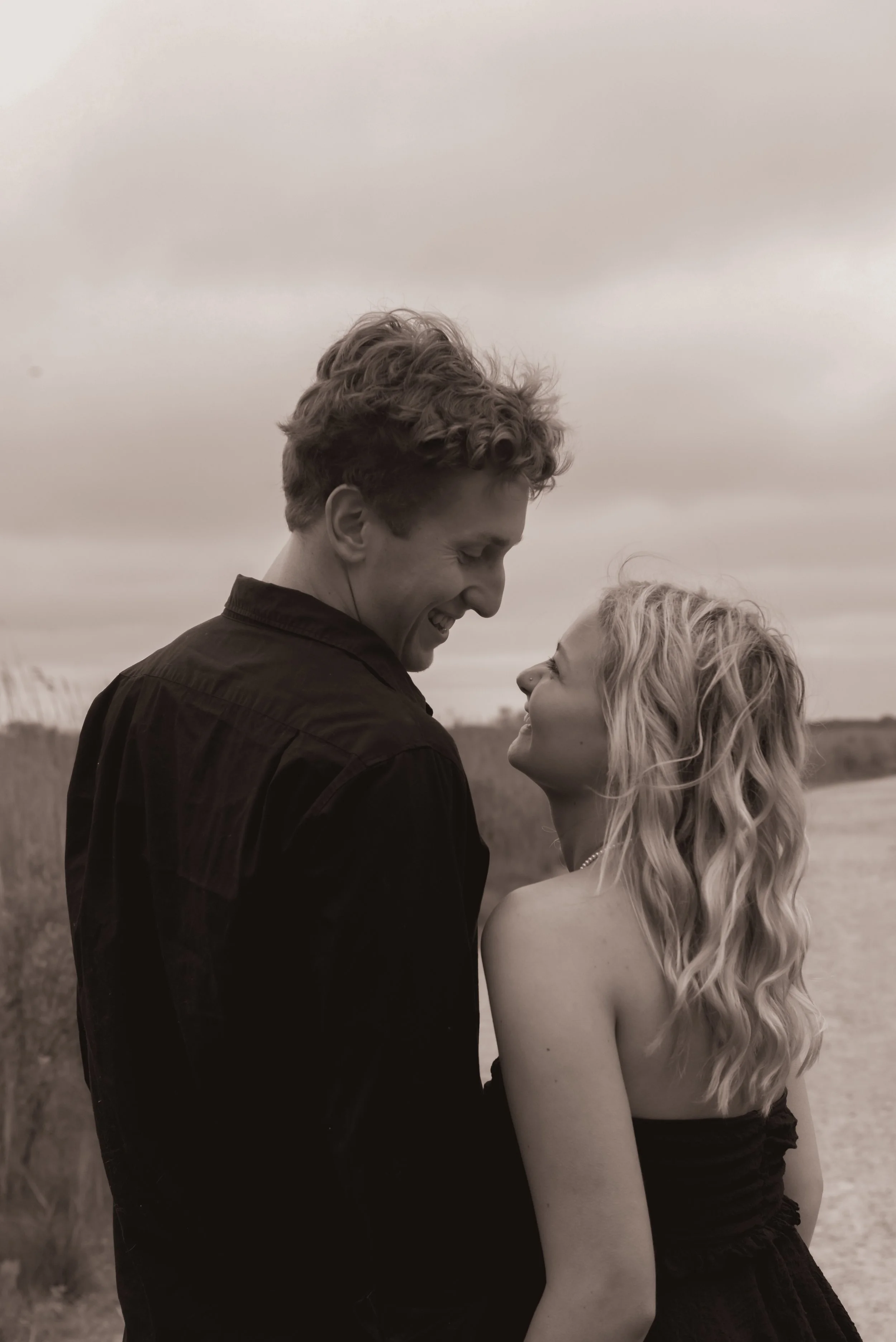 A smiling man and woman standing outdoors, looking at each other in a close embrace on a cloudy day.