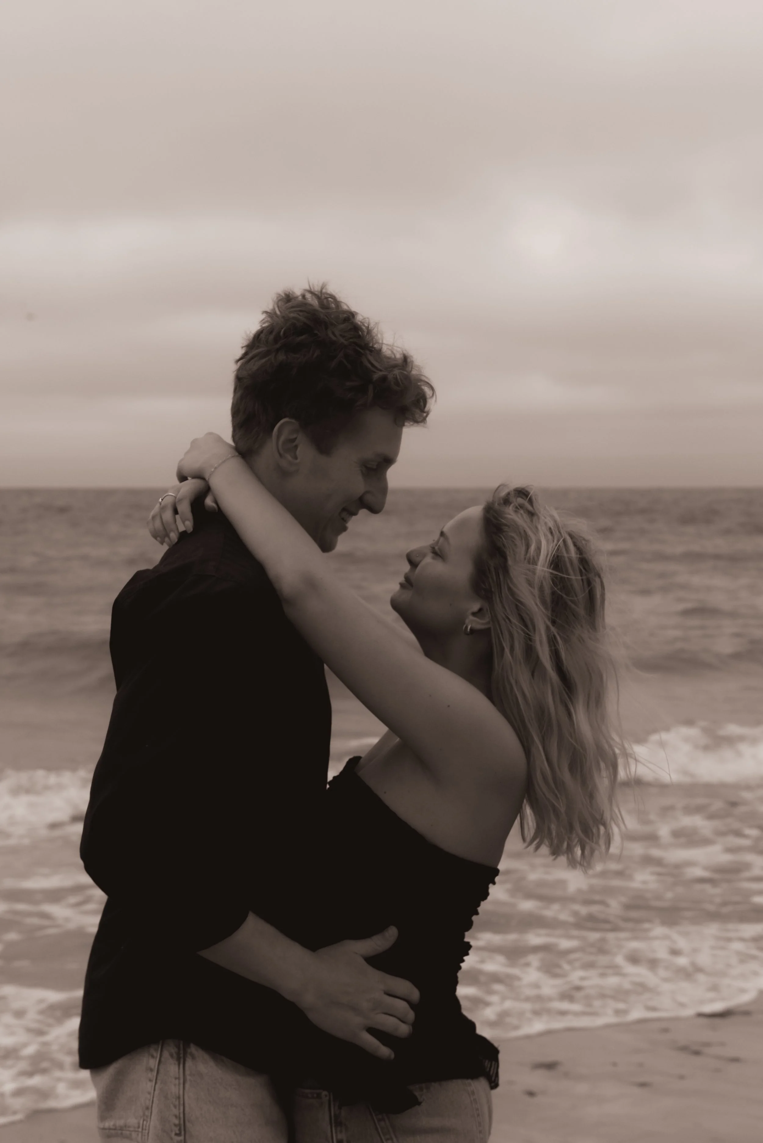 A couple sharing a romantic moment on the beach, embracing each other and smiling at each other with the ocean in the background.