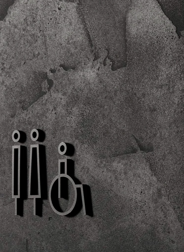 Metal restroom symbols for women, men, and wheelchair-accessible facilities on a textured black suavecrete wall by ambitec.