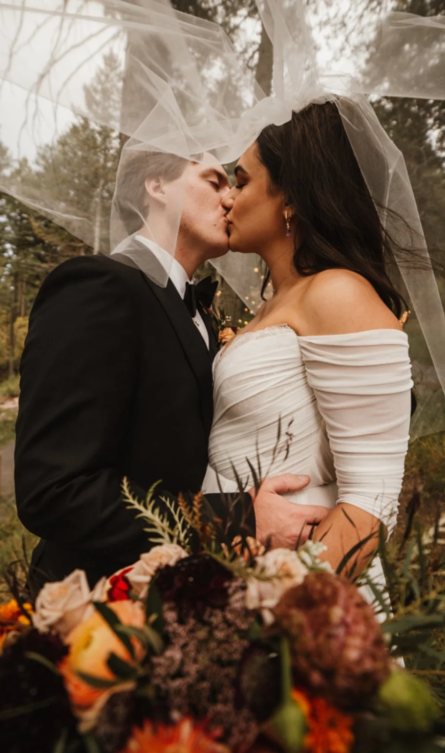 A newlywed couple is sharing a kiss under a veil outdoors, surrounded by trees and flowers.