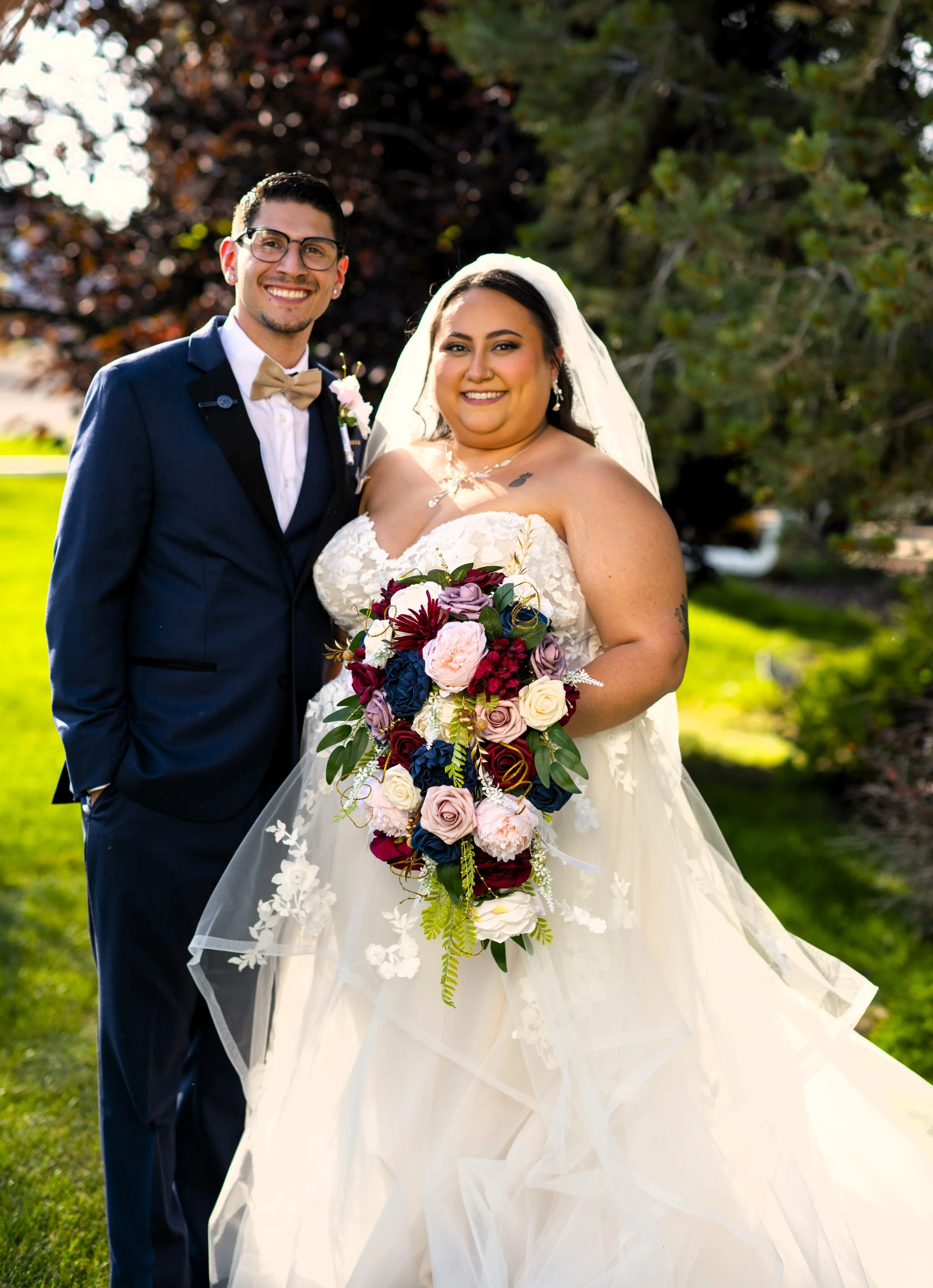A bride and groom outdoors on a sunny day, with trees and grass in the background, smiling and posing for a wedding photo.