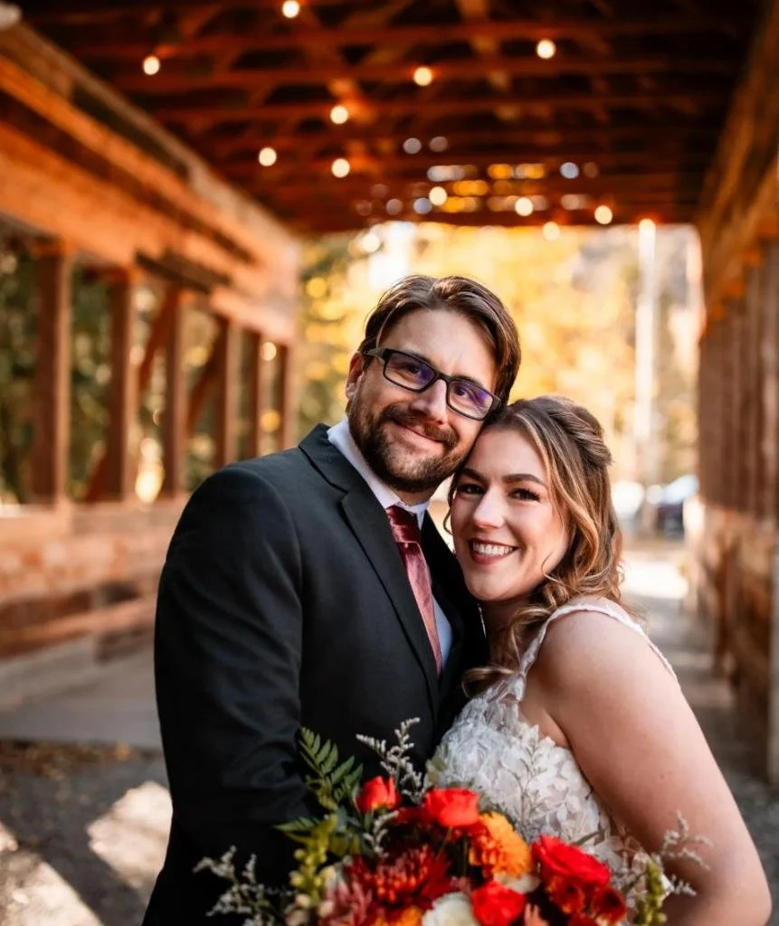 A newlywed couple smiling and embracing outdoors, with the bride holding a bouquet of red and orange flowers. The groom is wearing a black suit and glasses, and the bride is in a white lace wedding gown. The background shows a wooden structure and tr