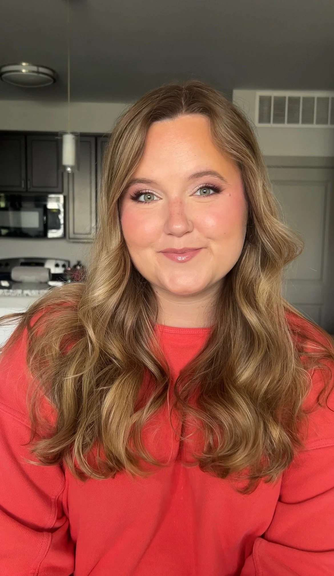 A woman with long, wavy light brown hair and gray eyes, wearing a red top, smiling at the camera, in a kitchen setting.