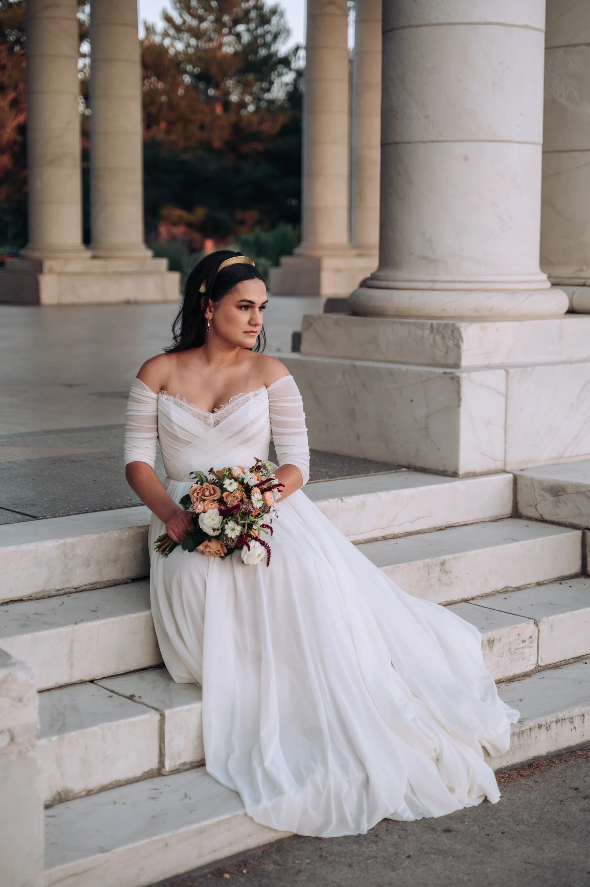 A bride in a white wedding dress sitting on marble steps holding a bouquet outside a classical building with large stone columns during sunset.