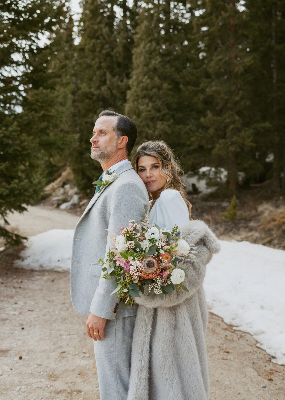 A couple dressed in wedding attire standing outdoors in a wooded area, with the woman holding a large bouquet of flowers and wearing a fur coat.