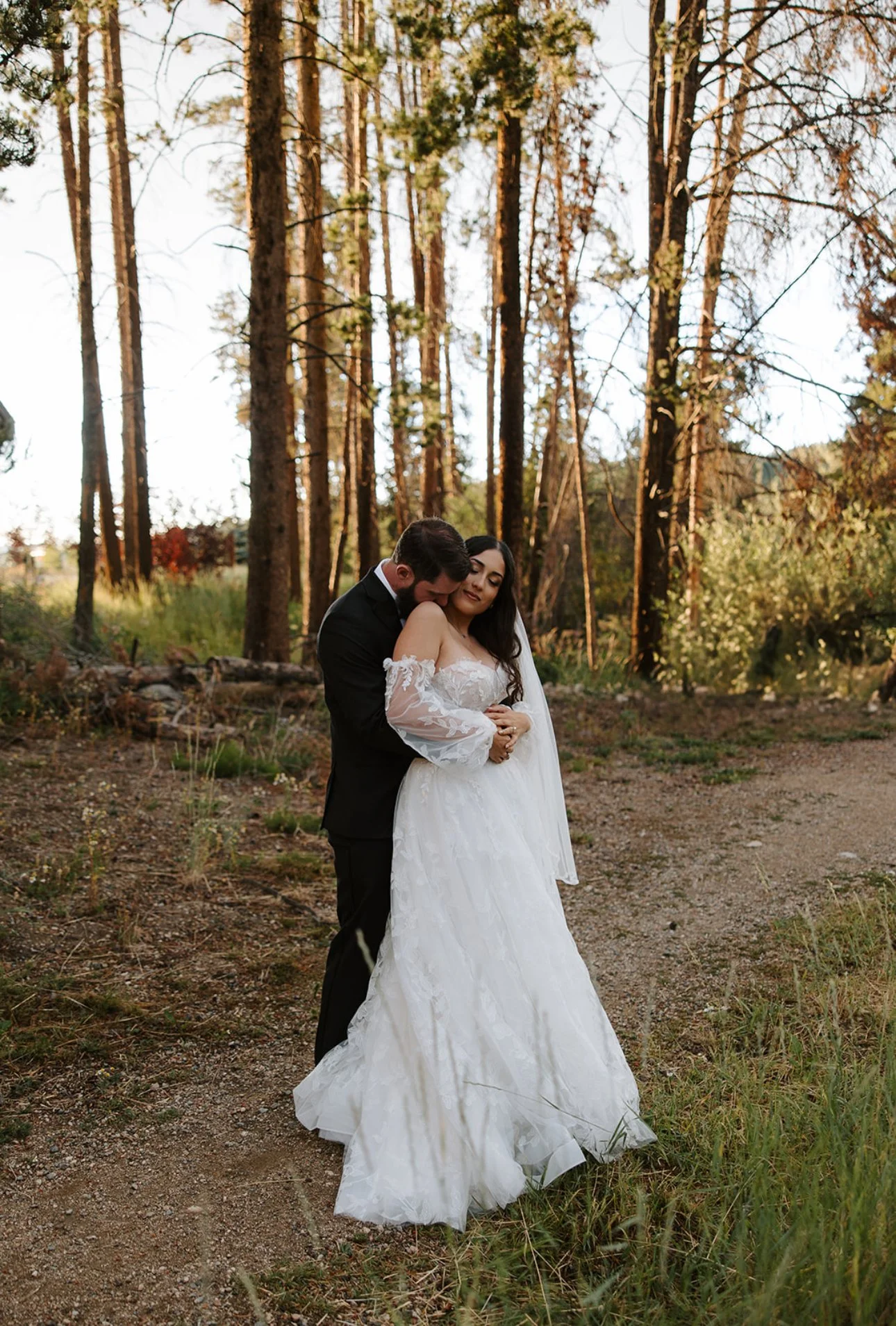 A bride and groom stand closely together outdoors in a wooded area during sunset, embracing each other. The bride wears a white wedding dress and the groom is in a black suit.