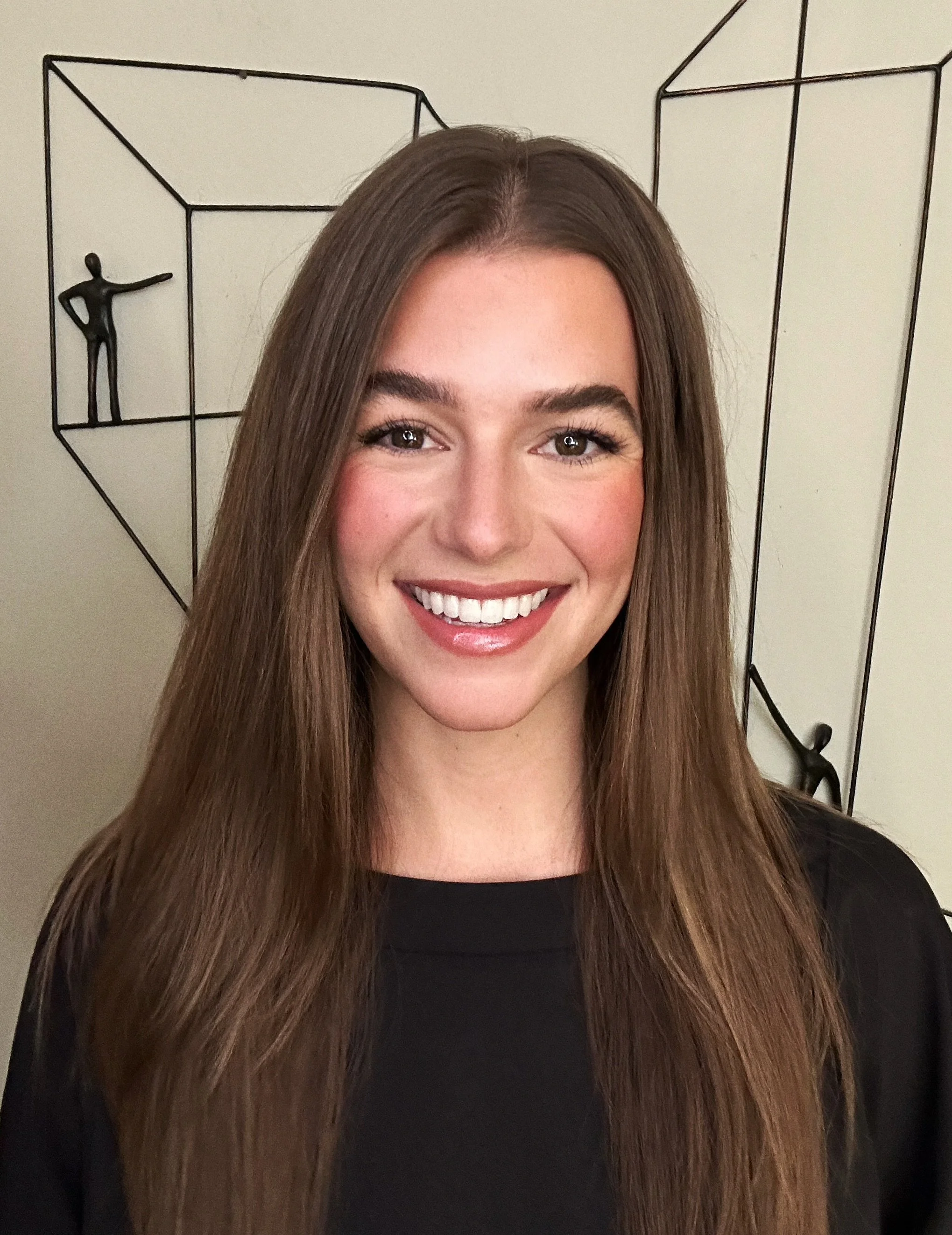 A woman with long brown hair smiling in front of a decorative wall art piece.