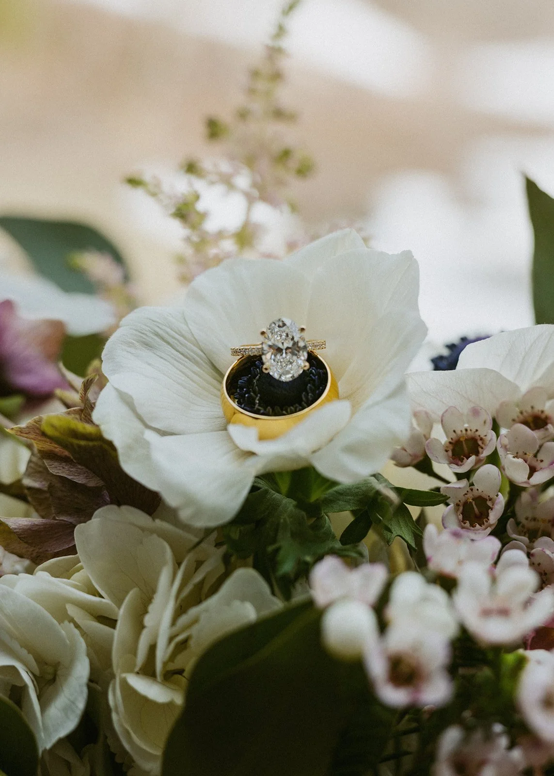 Close-up of a diamond engagement ring placed within a white flower, surrounded by various smaller flowers and greenery.