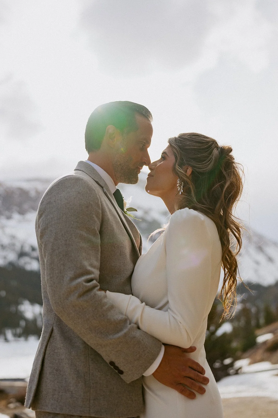 A bride and groom embrace outdoors during their wedding, standing close with their foreheads touching against a snowy mountain backdrop.