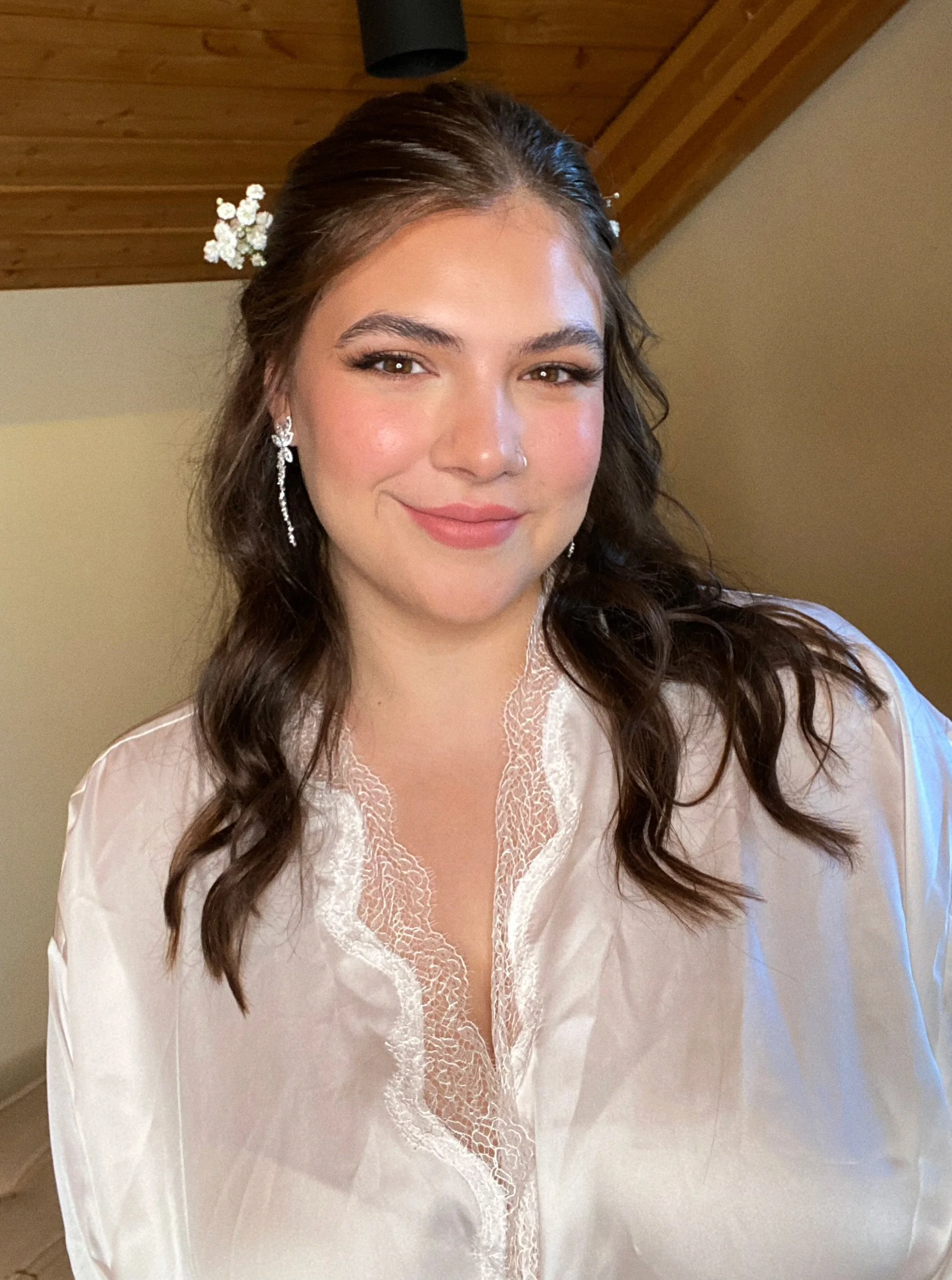 A young woman with long, dark wavy hair, wearing pearl and floral hair accessories, adorned with elegant earrings, and a lace-trimmed satin blouse, posing indoors with a wooden ceiling visible in the background.