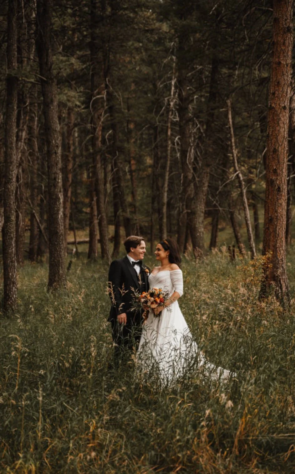 A newlywed couple stands in a forest, gazing at each other. The groom is dressed in a black tuxedo, and the bride is in a white wedding gown holding a bouquet of flowers.