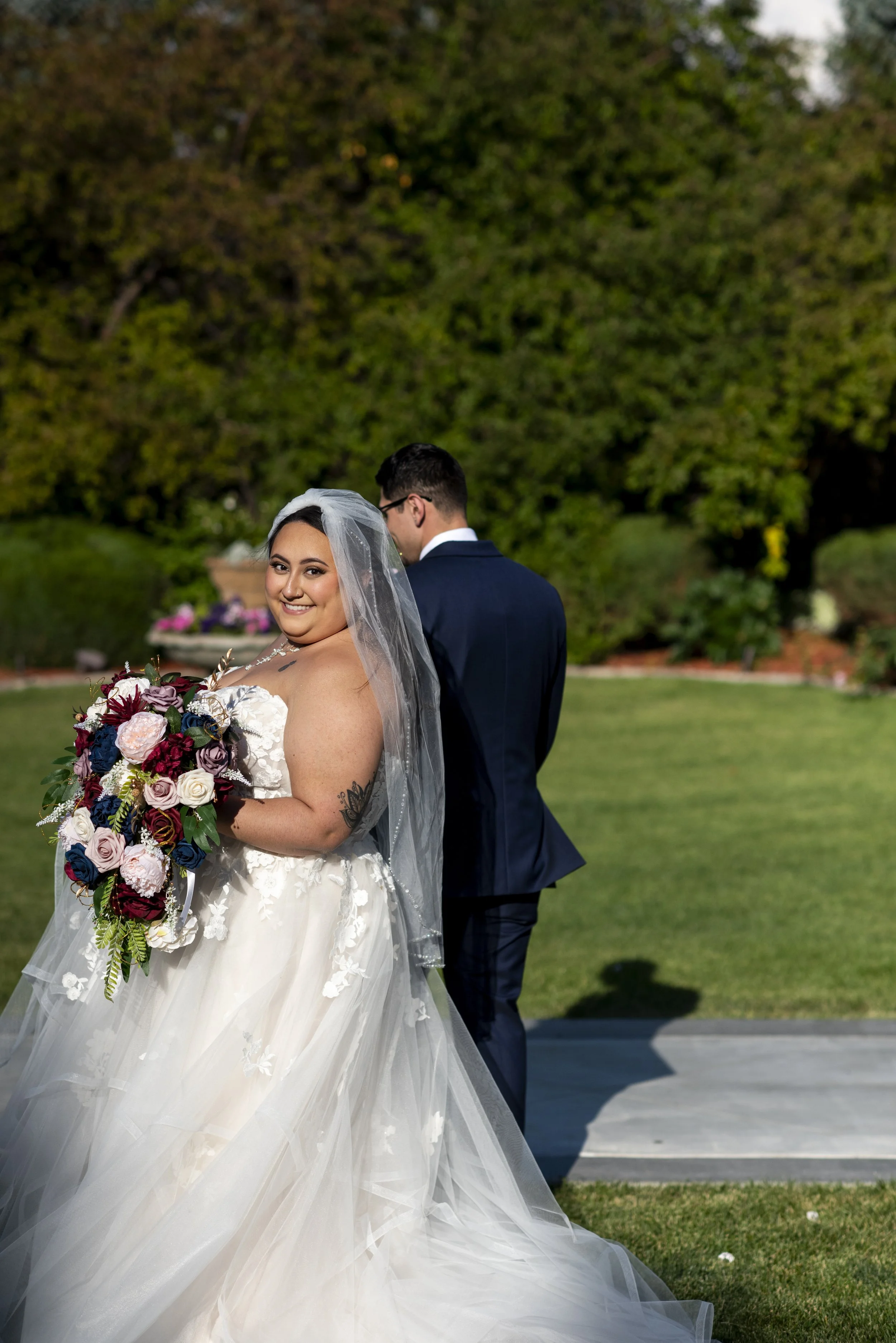 A bride in a white wedding gown holding a bouquet of flowers, smiling at the camera, with a groom in a dark suit standing behind her with his back to the camera in an outdoor setting with green trees and grass.