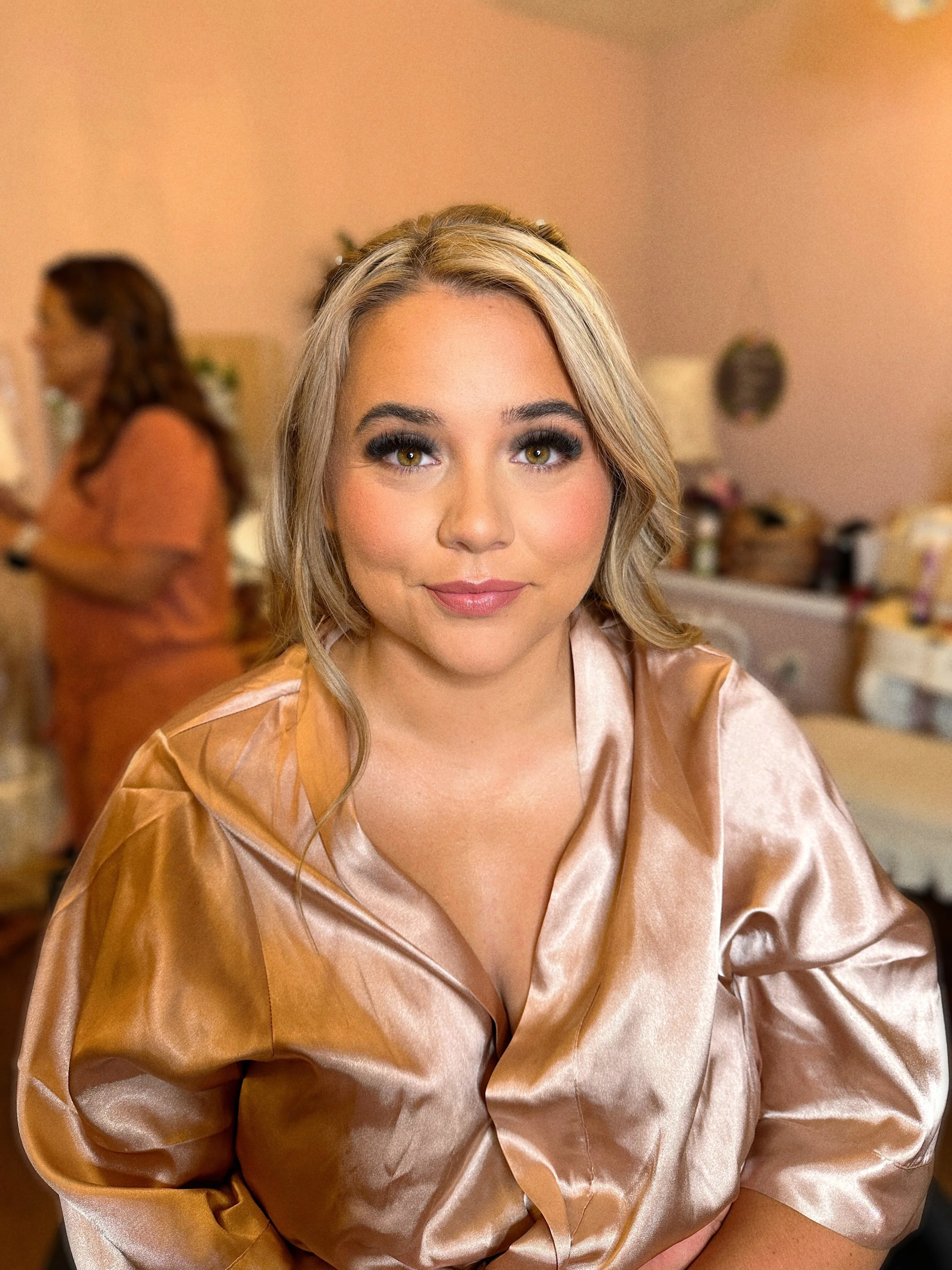 A woman with blonde hair and makeup wearing a satin tan-colored blouse, looking at the camera, with a blurred person in the background.