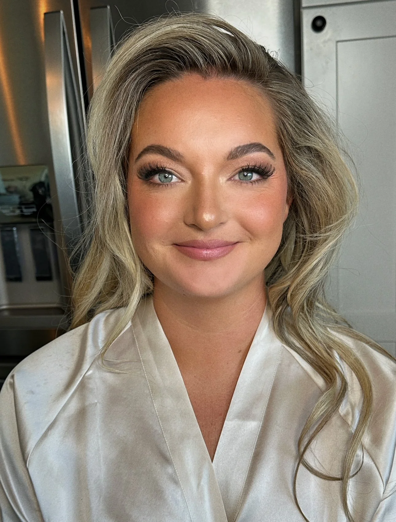 Close-up of a woman with blonde wavy hair, blue eyes, wearing makeup, and a beige satin robe, smiling in a modern kitchen.