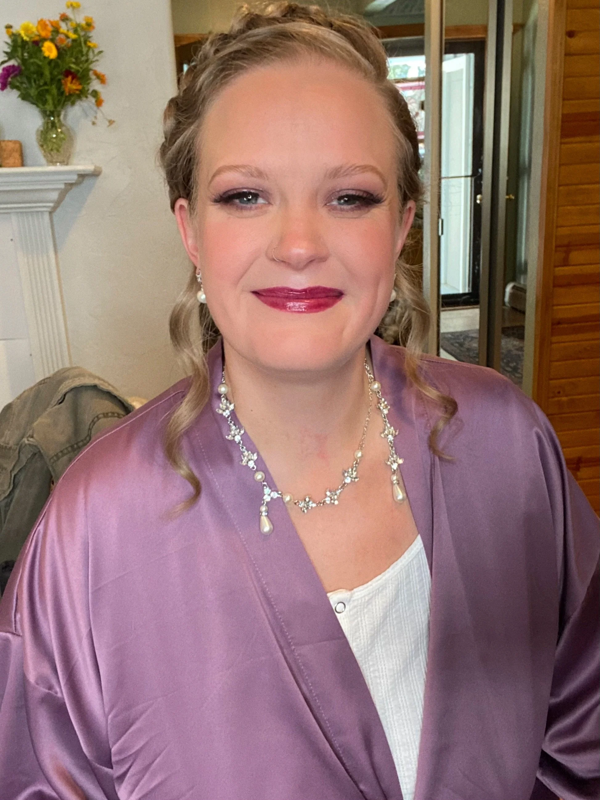 A woman wearing a lavender satin jacket and pearl jewelry, smiling indoors near a fireplace with a vase of flowers in the background.