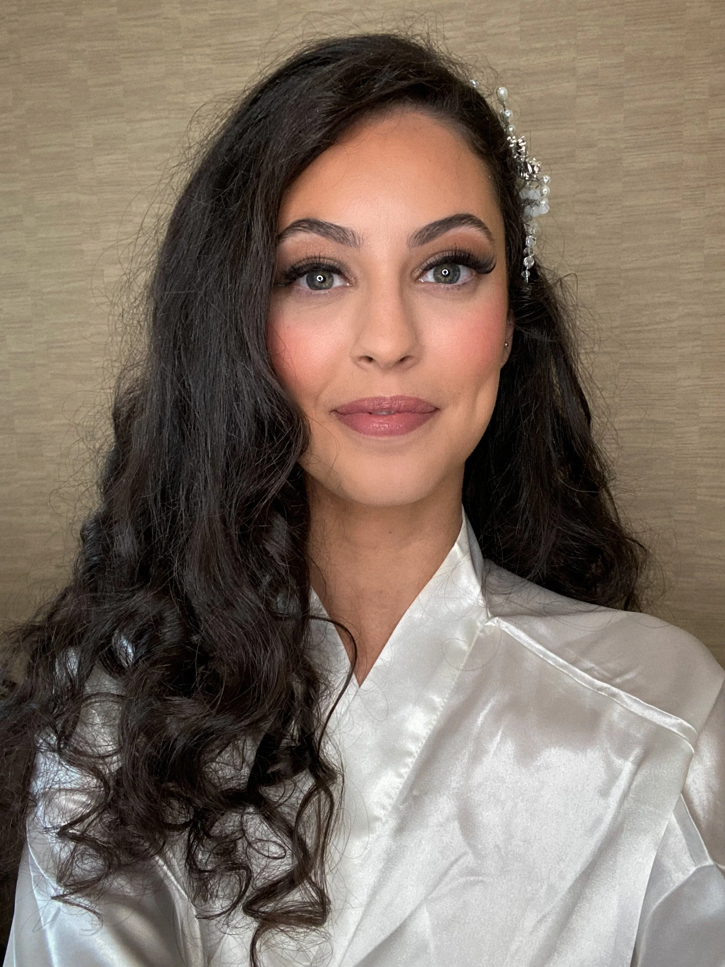 A woman with dark, wavy hair wearing a satin white garment and a decorative hair accessory with pearls, standing against a neutral background.