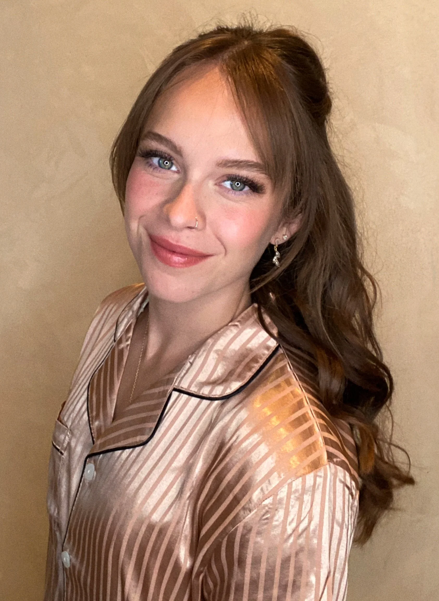 A young woman with long, wavy brown hair and blue eyes, wearing a satin striped pajama top with a collar and buttons, standing against a neutral background.