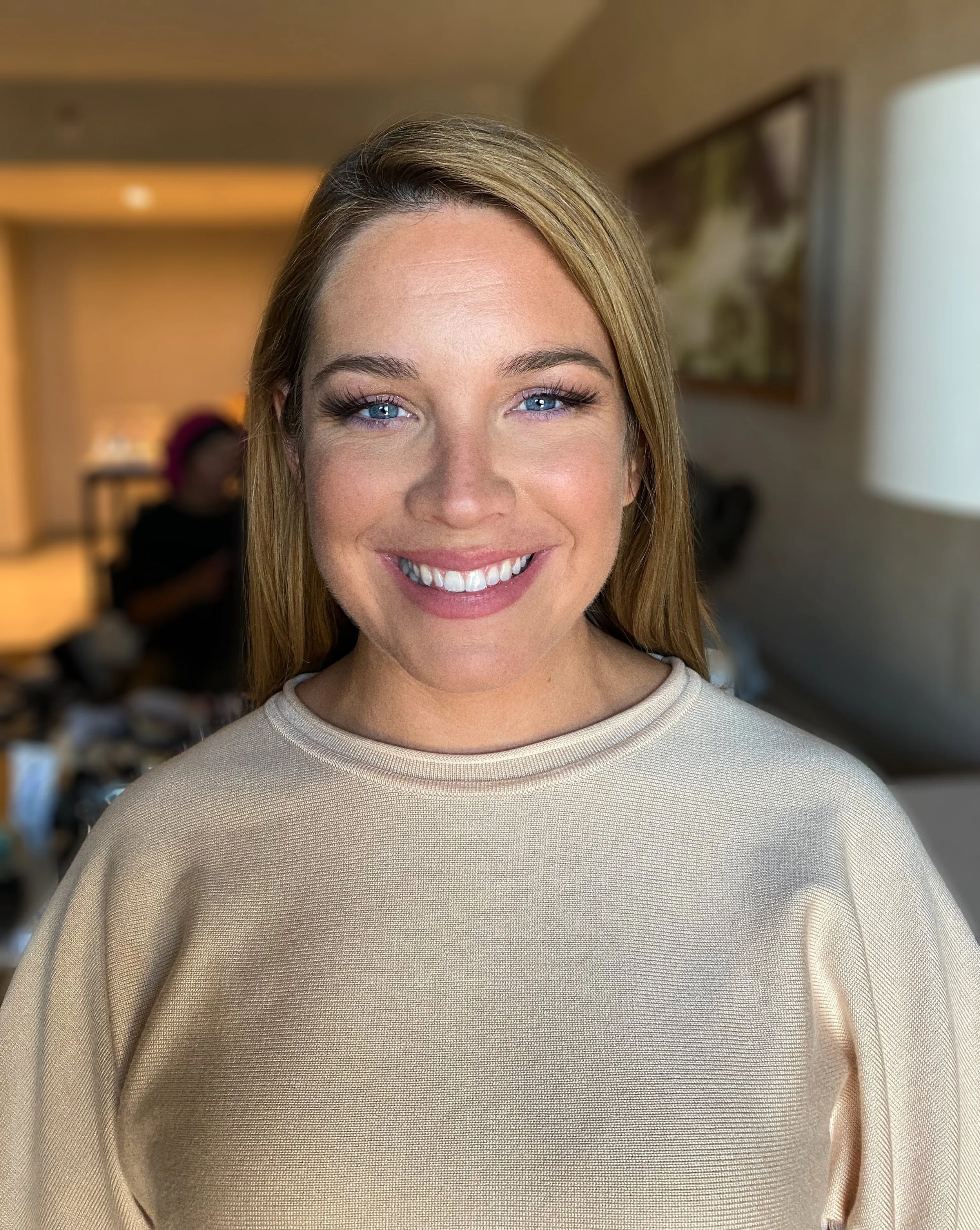 A smiling woman with light brown hair, blue eyes, and wearing a beige sweatshirt, standing indoors with a blurred background.