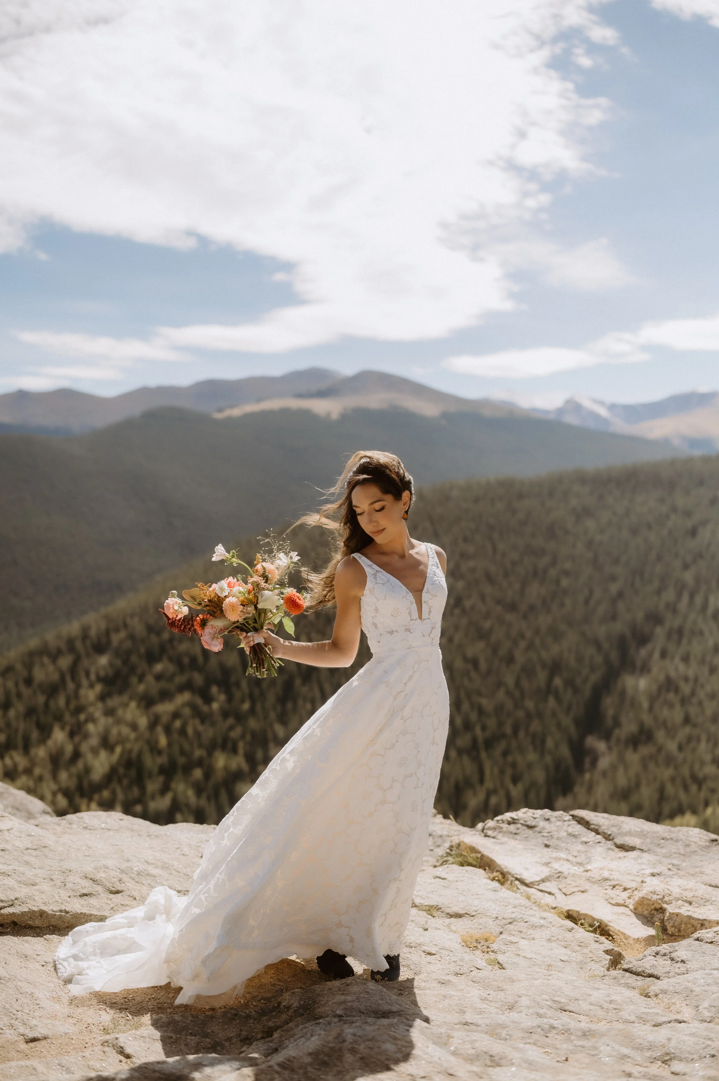 A woman in a white wedding dress holding a bouquet of flowers outdoors on a rocky ledge with mountains and a partly cloudy sky in the background.