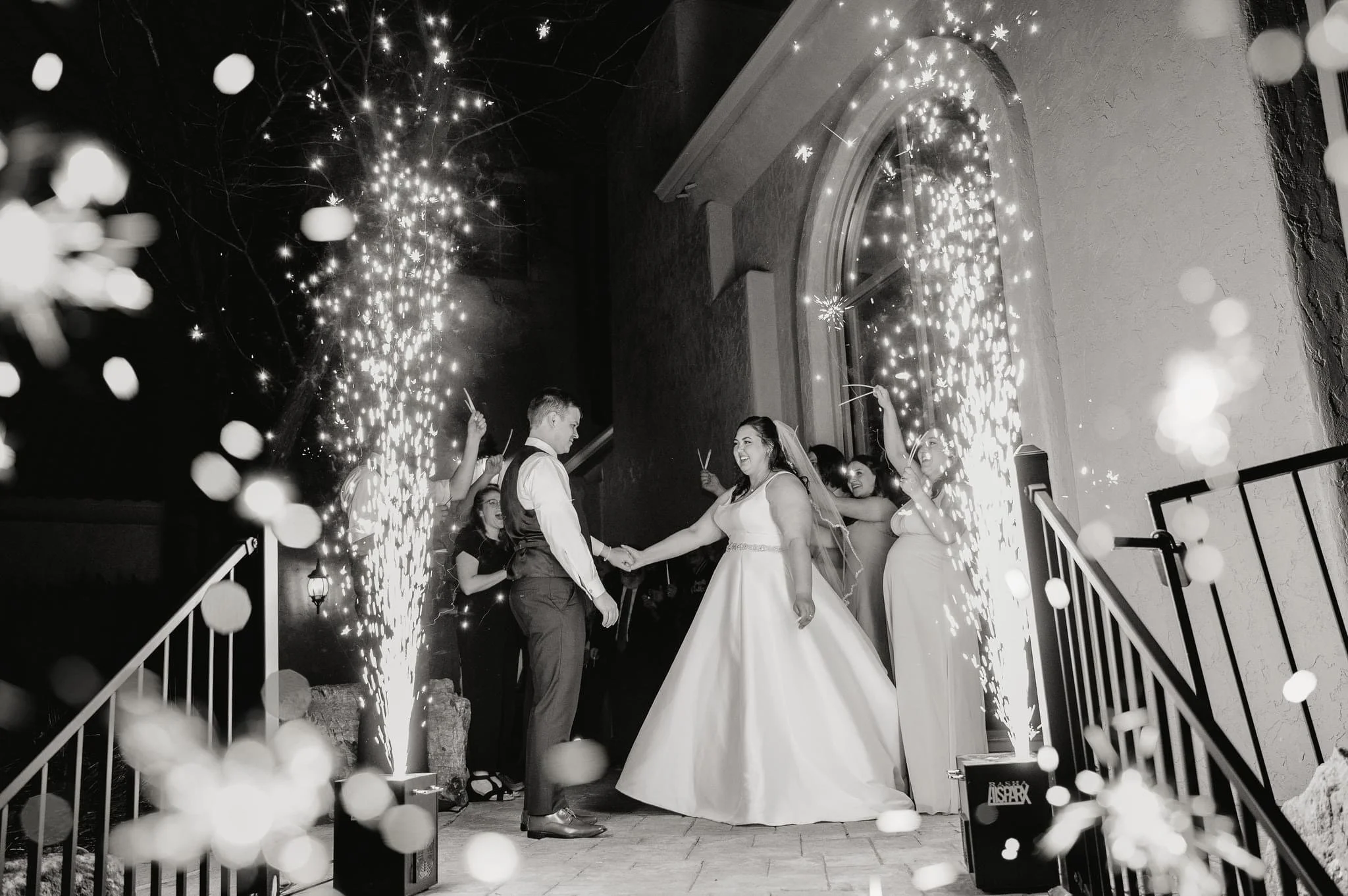 A bride and groom holding hands and smiling at each other during their wedding reception outside at night. They are surrounded by friends or family celebrating with sparklers that are creating bright, sparkling arcs in the air. The scene is festive a