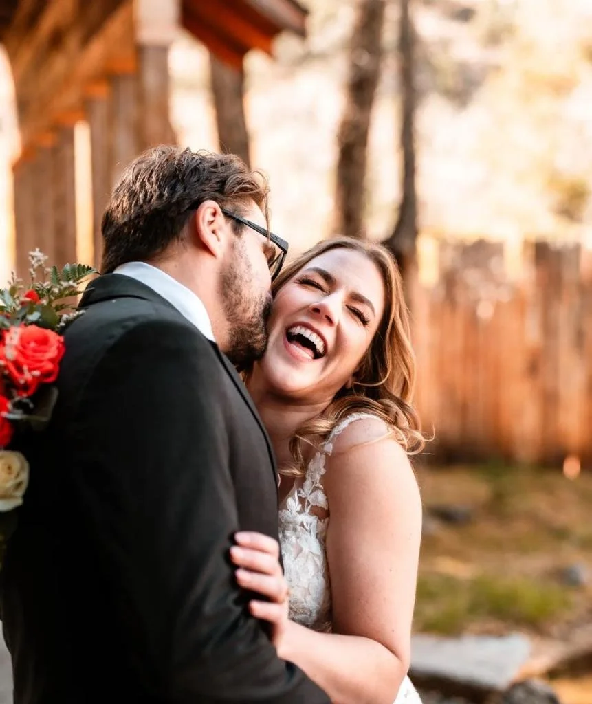 A couple sharing a joyful moment outdoors, with the woman smiling and the man holding a bouquet of flowers, in a wooded area.
