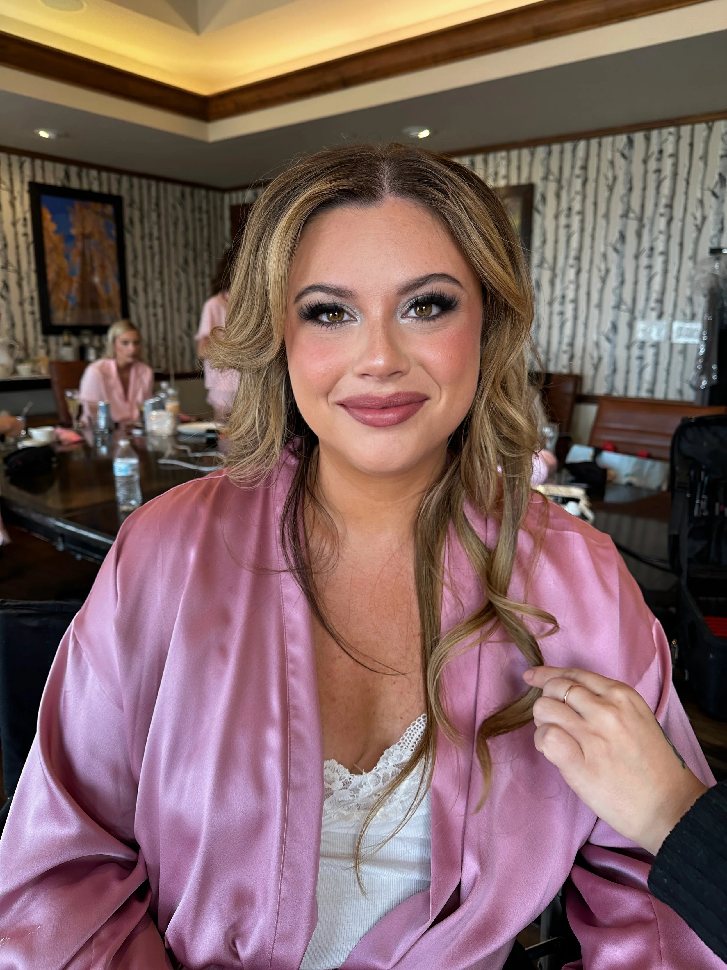 A woman with styled hair and makeup in a pink satin robe, smiling at the camera in a room with people preparing, surrounded by hair and makeup tools.