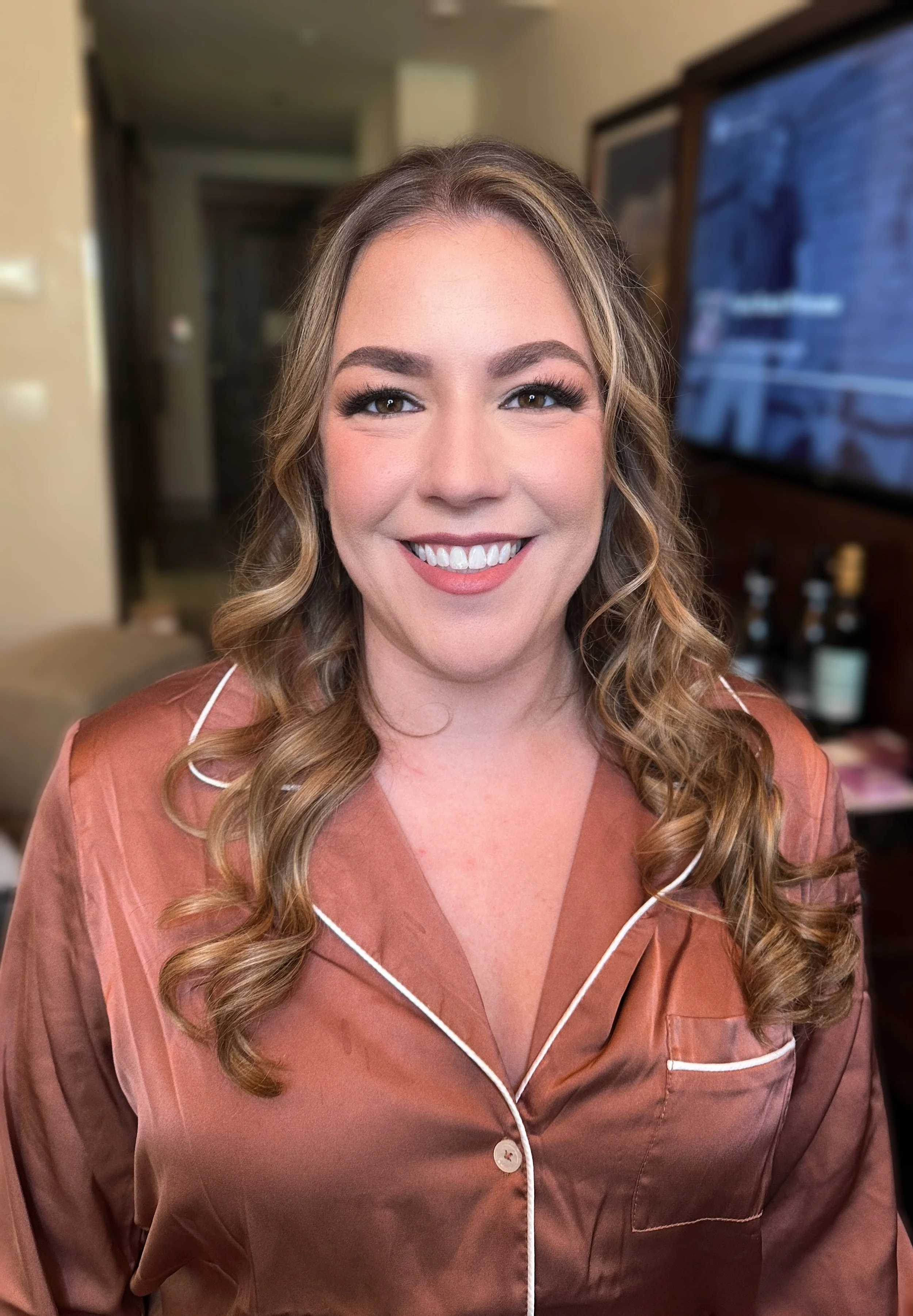 A woman with long curly hair, wearing brown satin pajamas with white piping, smiling in a room with a large TV behind her and a beige wall.