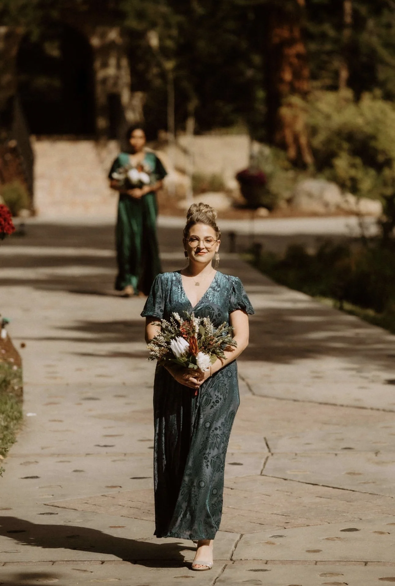 A woman in a teal dress holding a bouquet of flowers walking on a stone pathway outdoors, with another woman in the background holding flowers.
