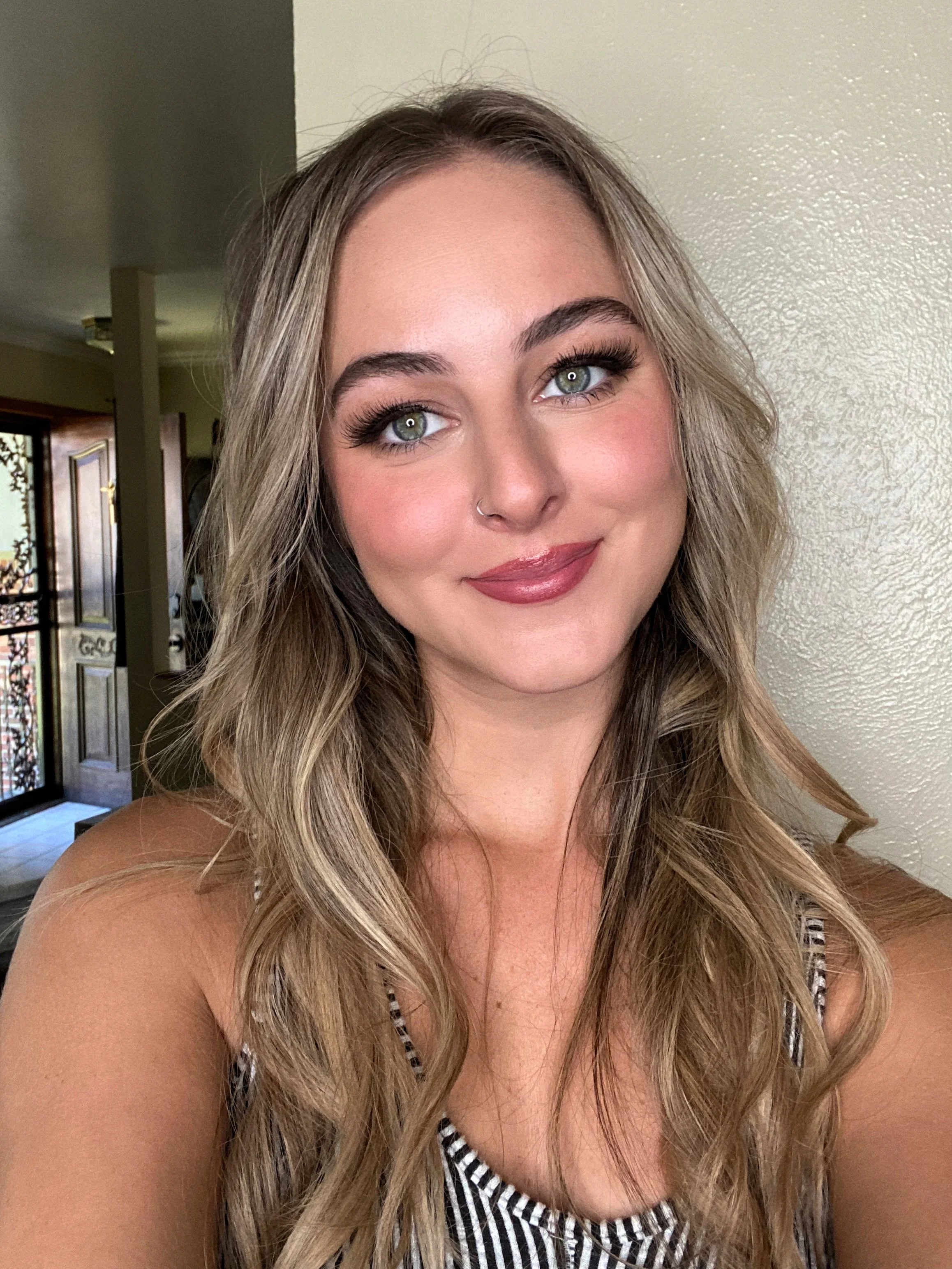 Close-up of a young woman with blonde hair, green eyes, and a nose ring, smiling at the camera, indoors.