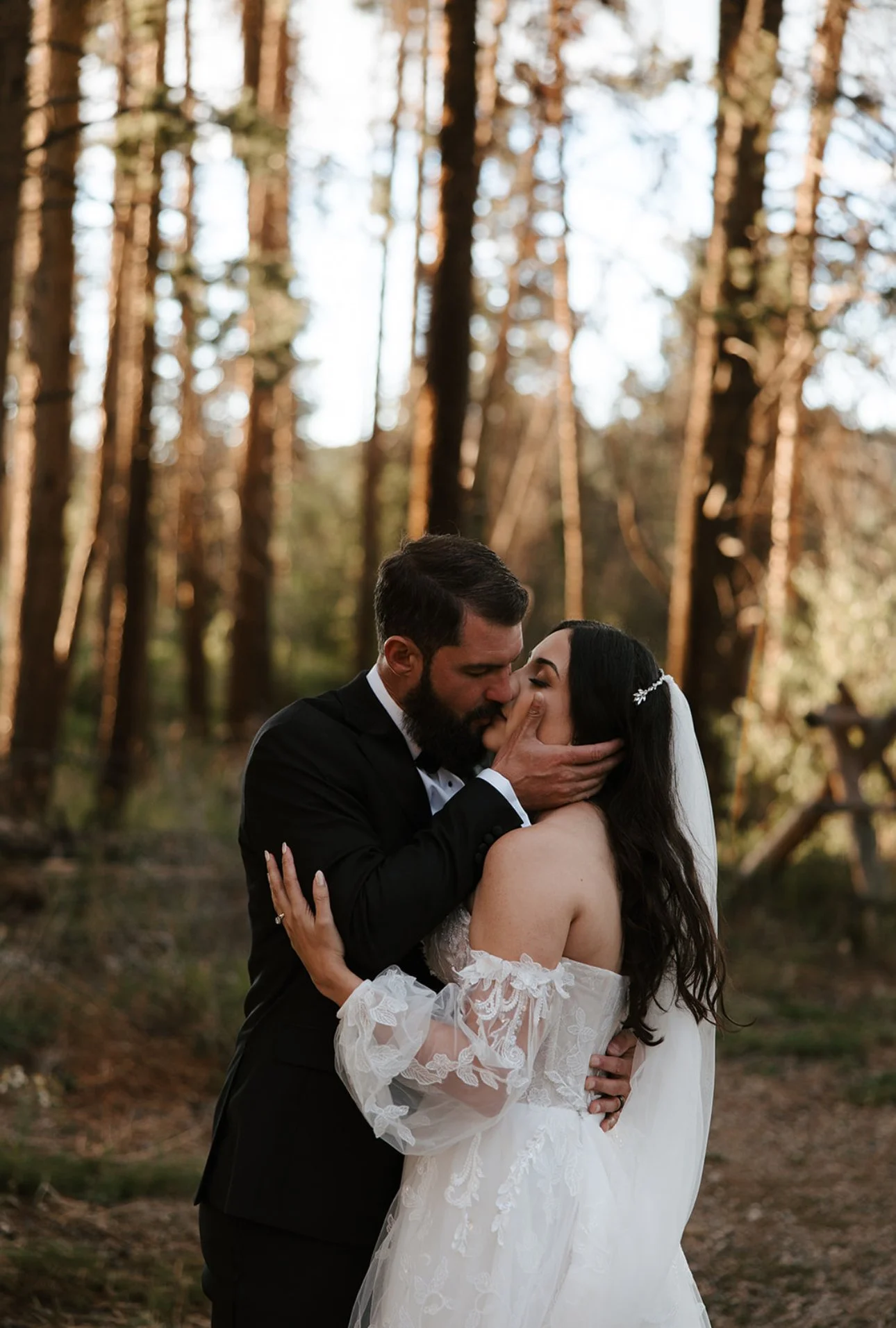 A couple sharing a kiss in a forest during sunset, with tall trees in the background. The man is in a black suit, and the woman is in a white wedding dress with off-the-shoulder lace sleeves.