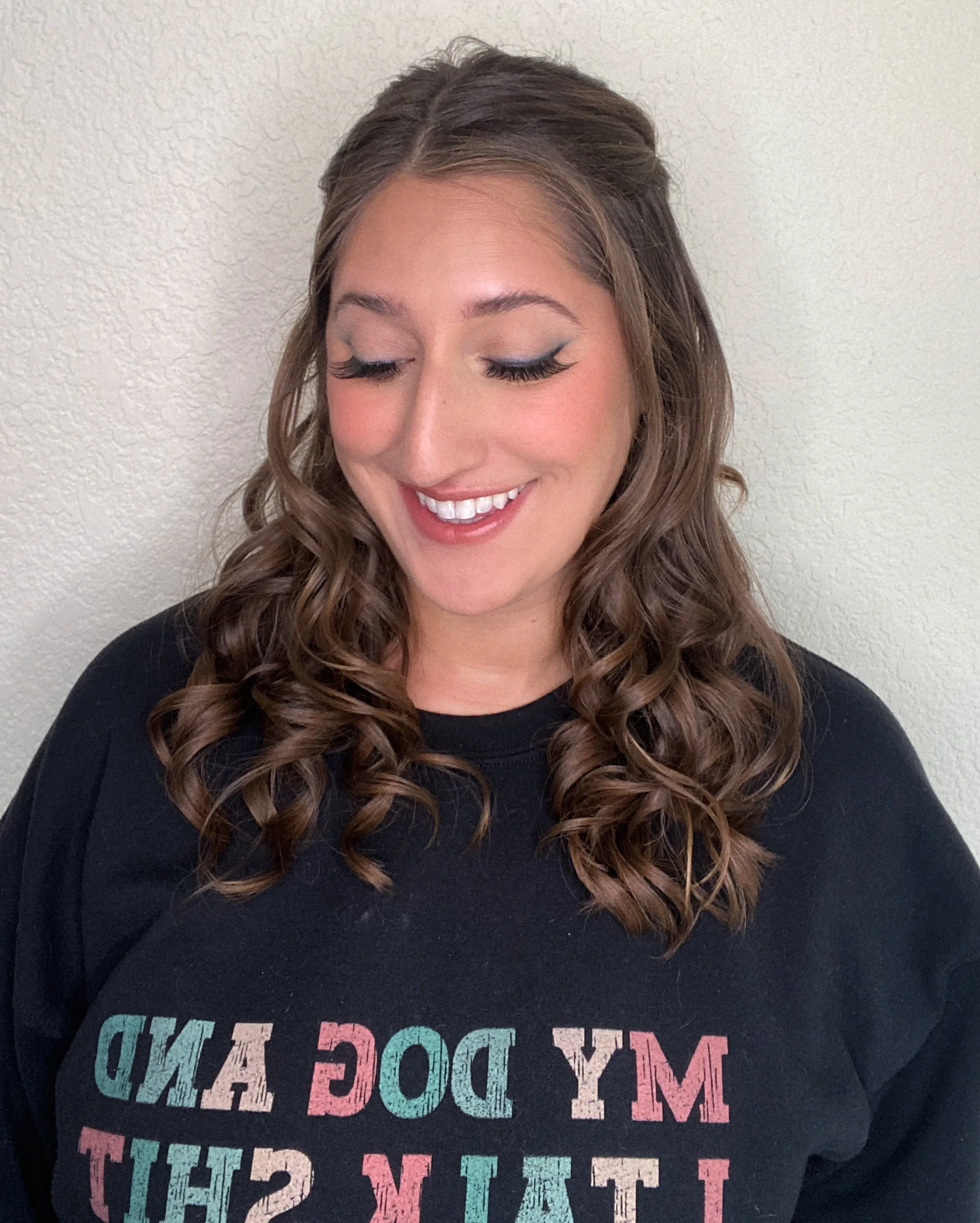 A woman with wavy brown hair, smiling and looking down, wearing a black t-shirt with colorful text.