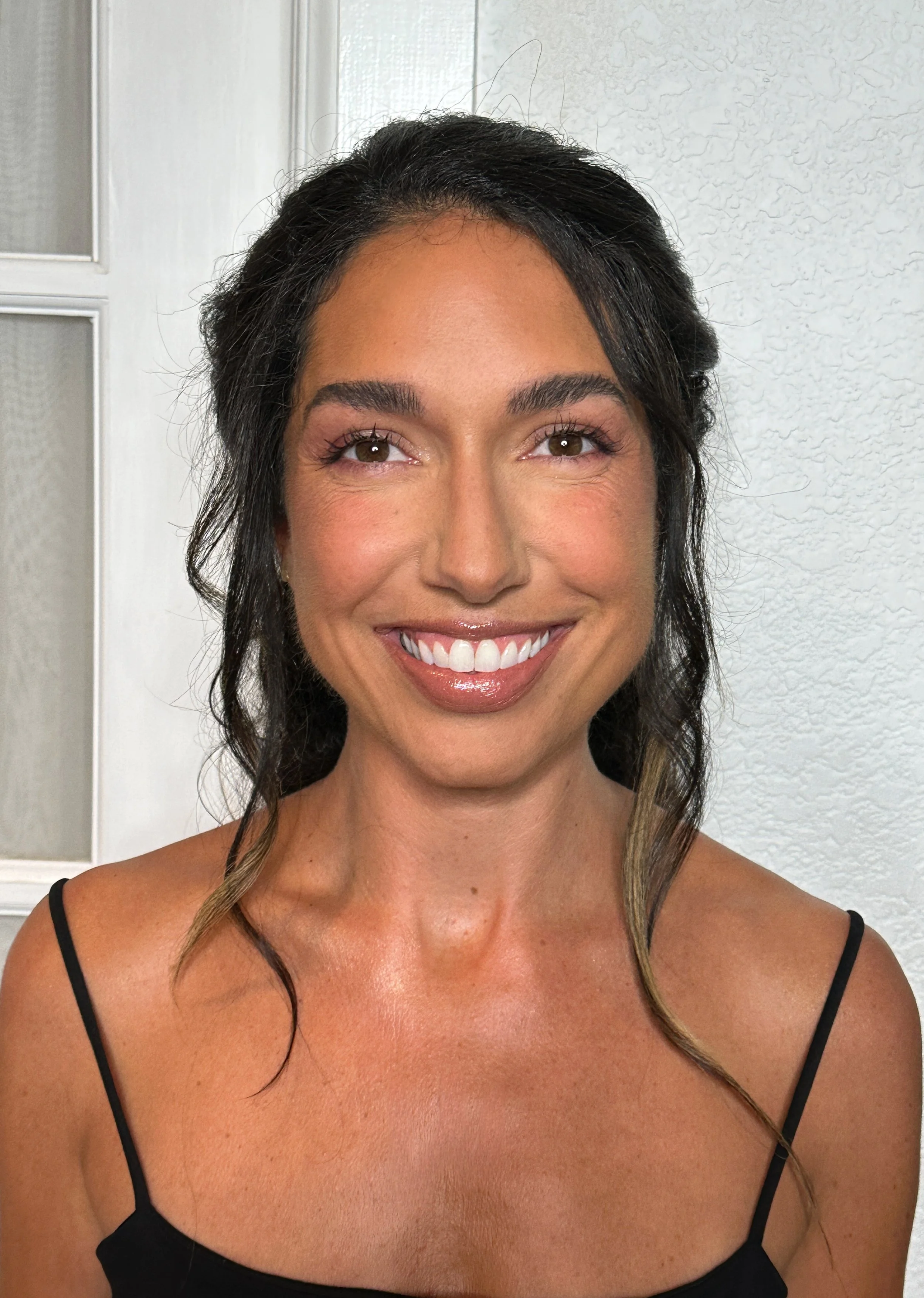 A woman with dark hair, wearing a black spaghetti strap top, smiling in front of a white textured wall and window.