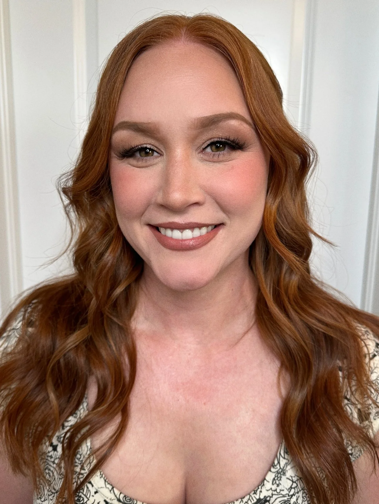 A smiling woman with long wavy red hair, wearing makeup, and a patterned top, standing in front of a white door.