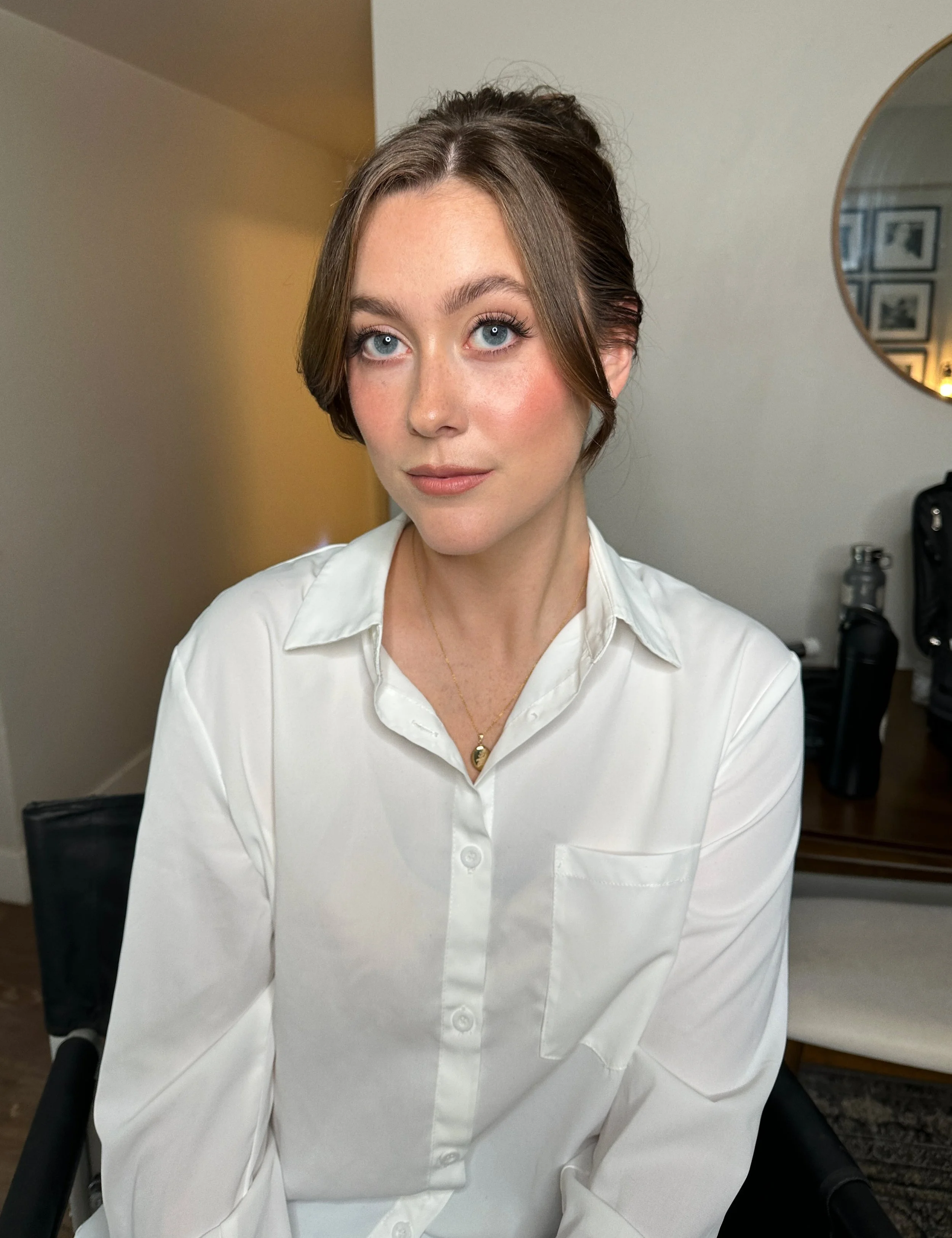 A woman with brown hair styled up, wearing a white button-up shirt and a gold necklace, sitting indoors in front of a neutral wall and a round mirror.