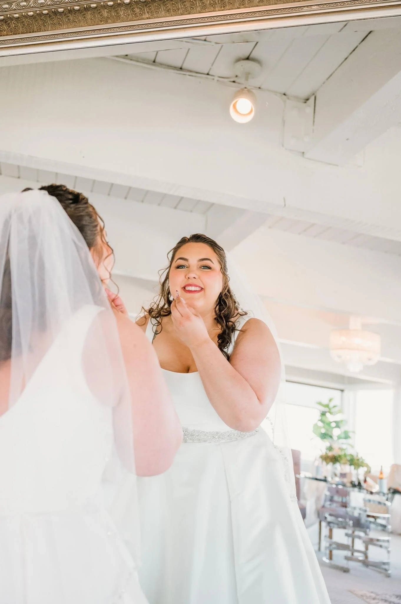 Bride with veil looking in mirror as friend or makeup artist applies makeup in a brightly lit room decorated with plants and modern furniture.