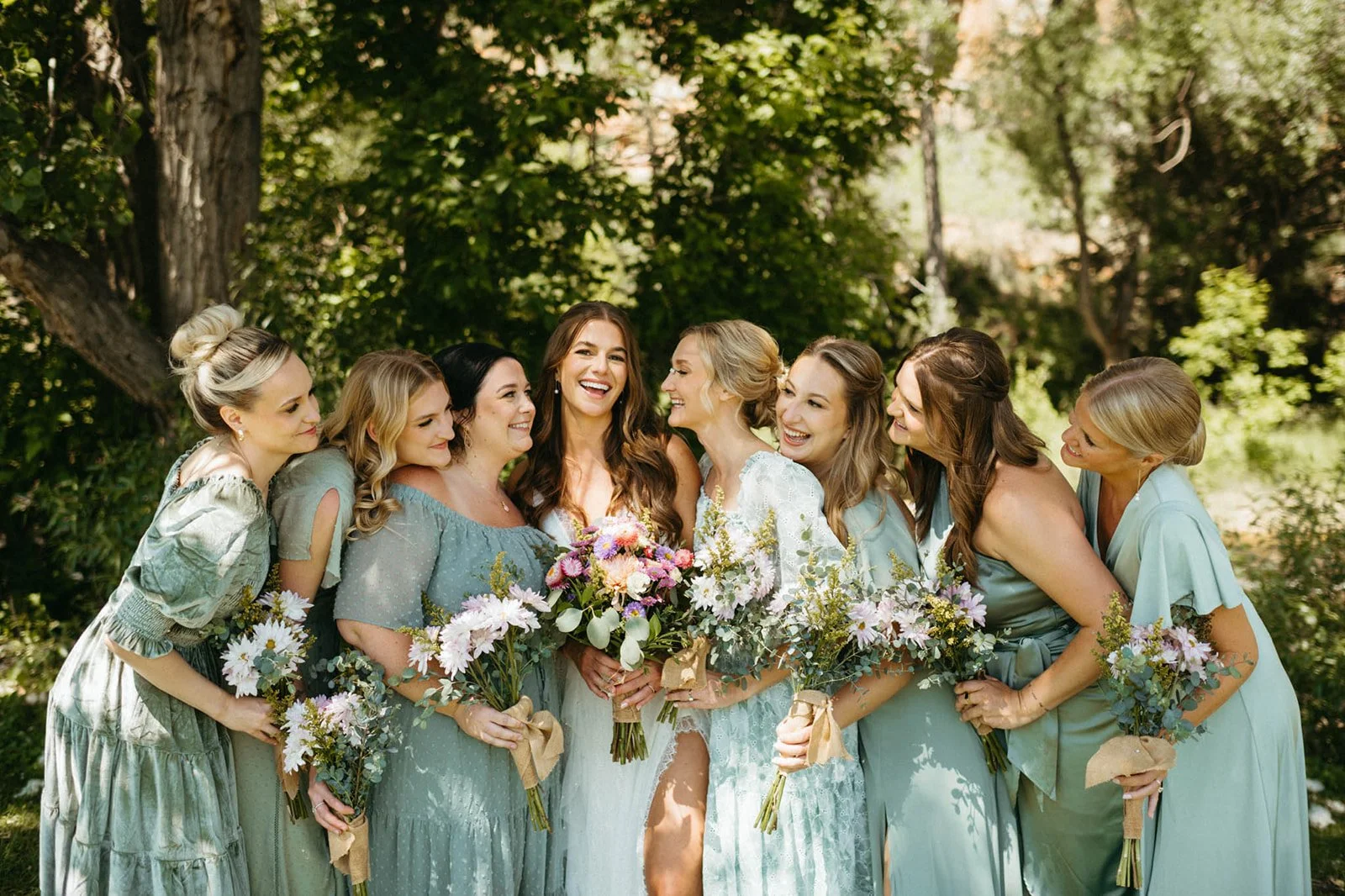 A group of seven women, including a bride in a white dress, smiling and holding bouquets, standing outdoors in a lush green park with trees in the background.
