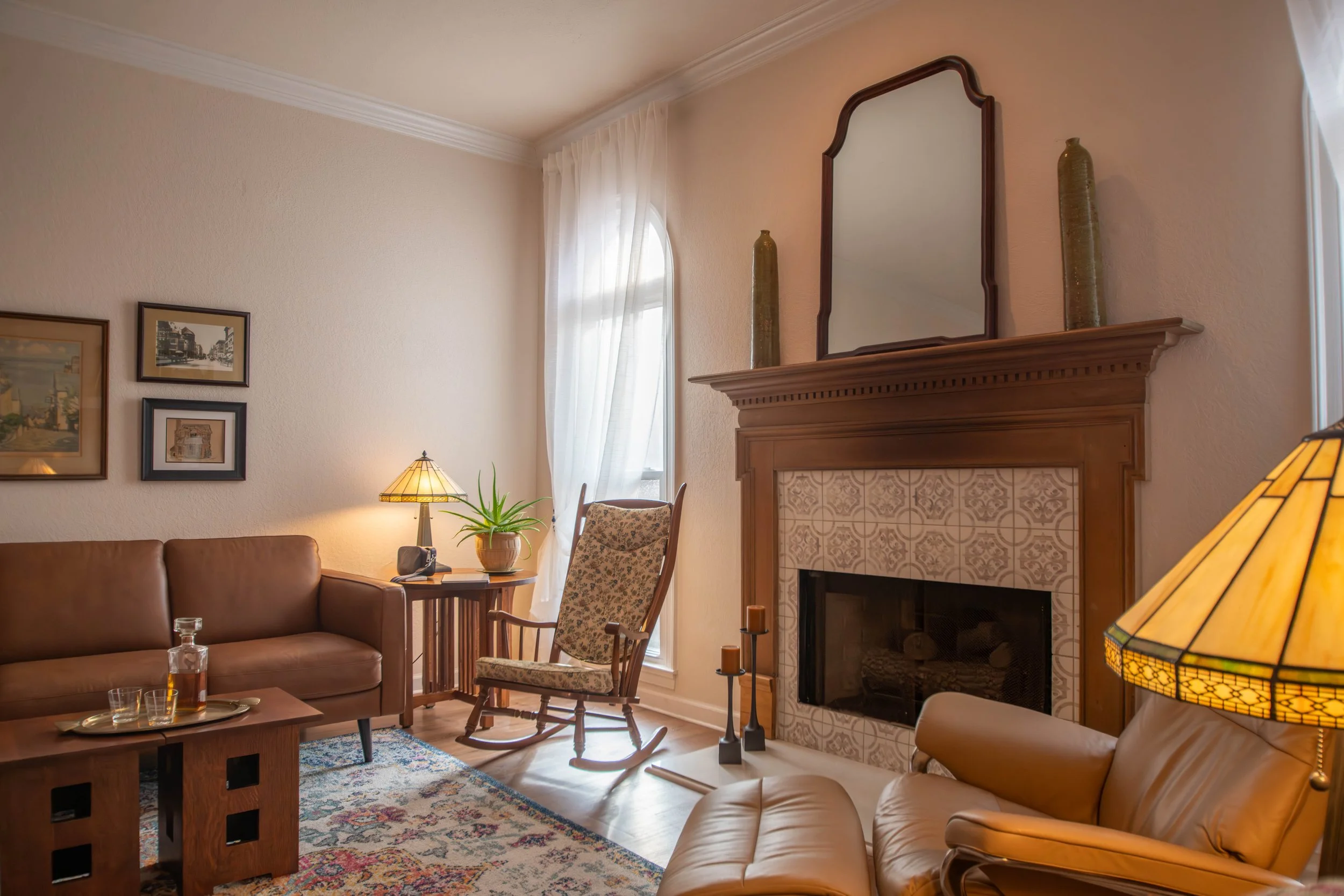 Warm traditional living room featuring a wooden fireplace mantel, rocking chair, leather furniture, side table with whiskey glasses, and soft natural light from window – Mac Schaaf interior photography