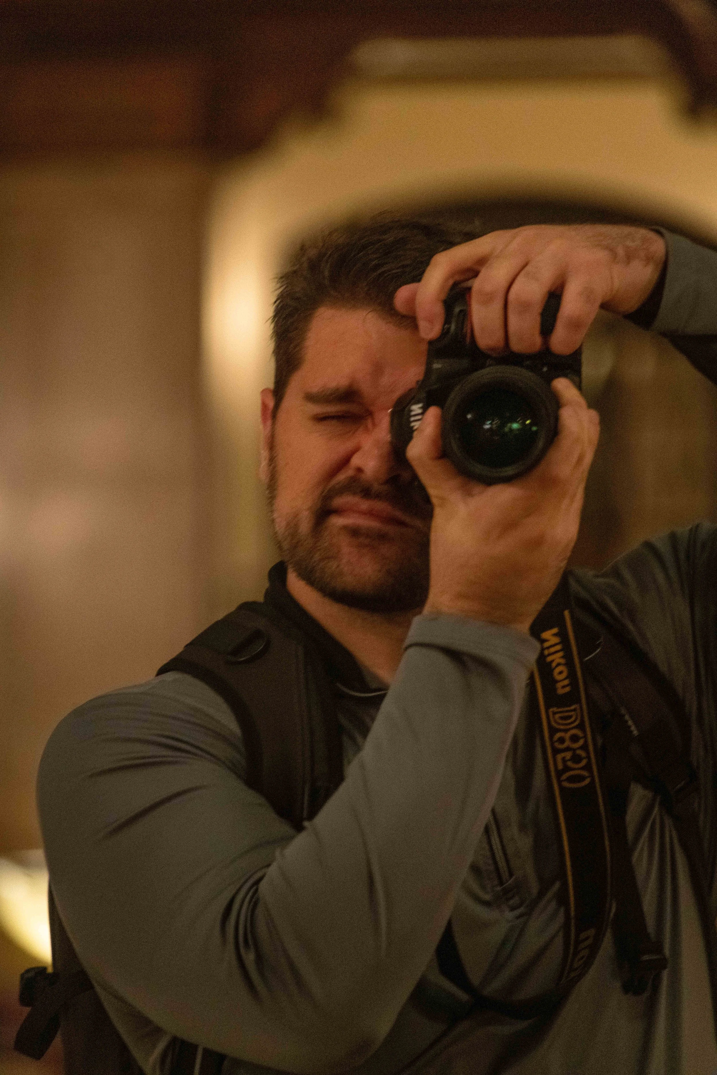 A man with dark hair, a beard, and a serious expression is taking a photo with a professional Nikon camera. He is wearing a gray and black jacket and carrying a backpack, standing indoors with a blurred background.