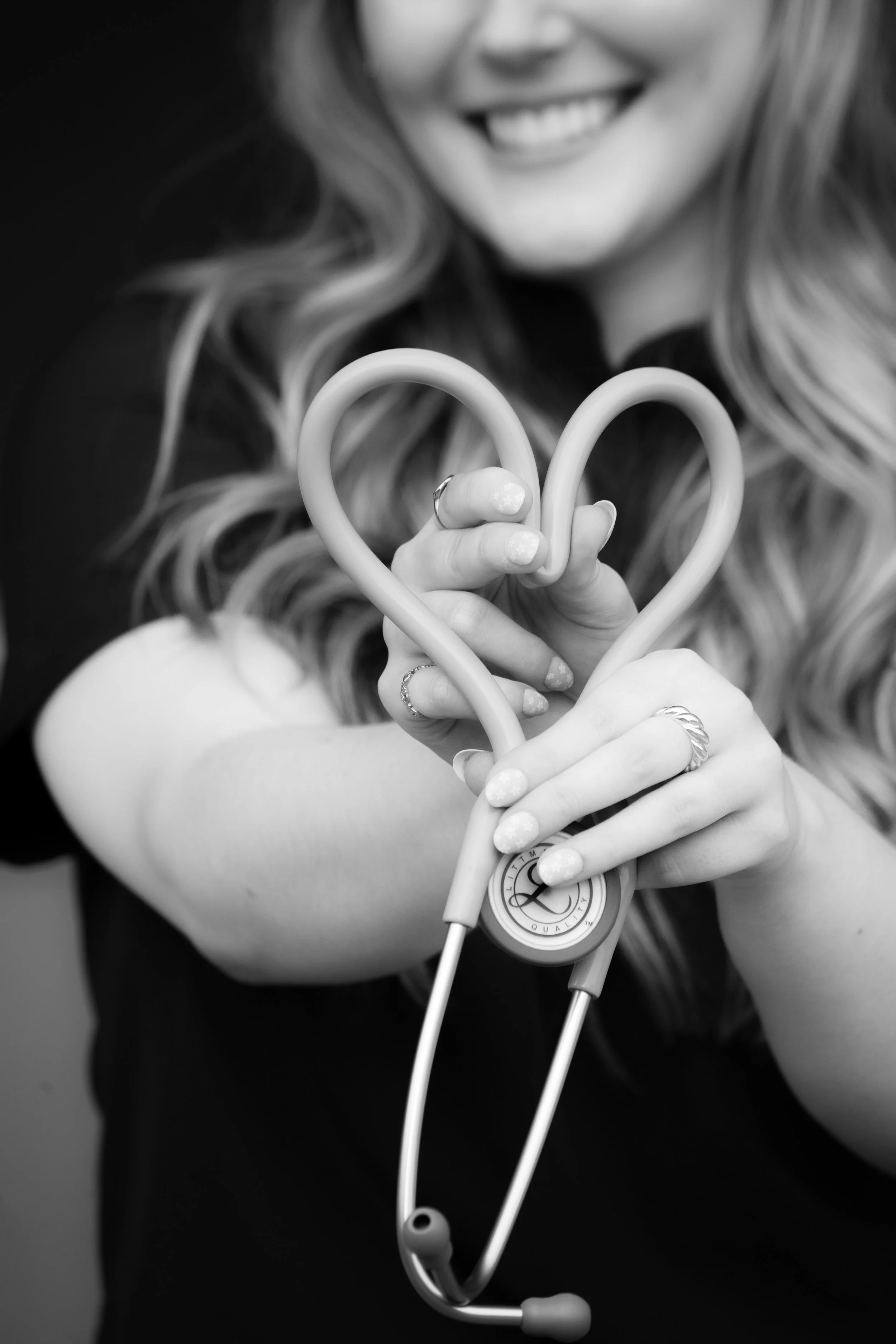 Black and white portrait of a smiling nurse holding a stethoscope shaped like a heart, close-up on hands and face, medical professional showing care and compassion