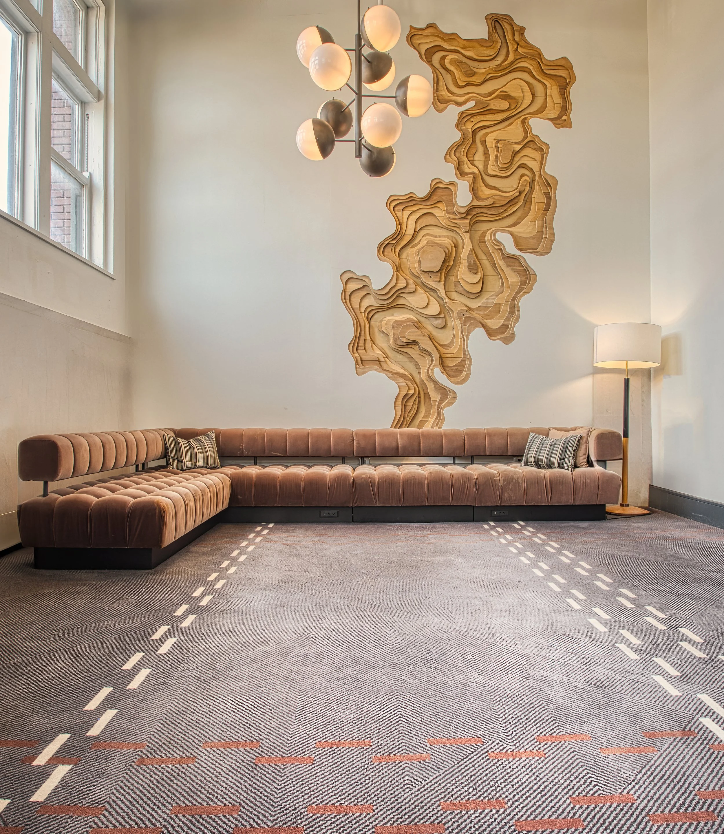 Modern living room with a long brown velvet sectional sofa, a large abstract wooden wall art piece, a tall lamp, and a spherical chandelier, with a window on the left side.