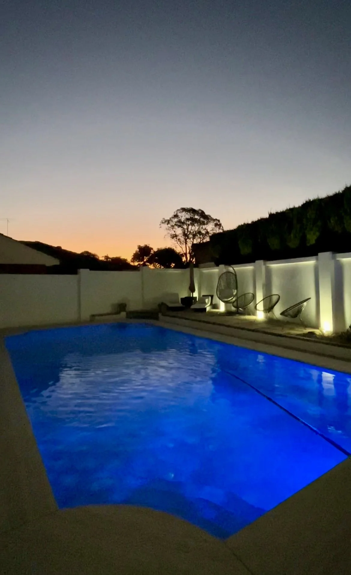 A backyard swimming pool illuminated with blue lights at dusk, with a white privacy fence and lounge chairs along the deck, and a sunset sky with trees in the background.