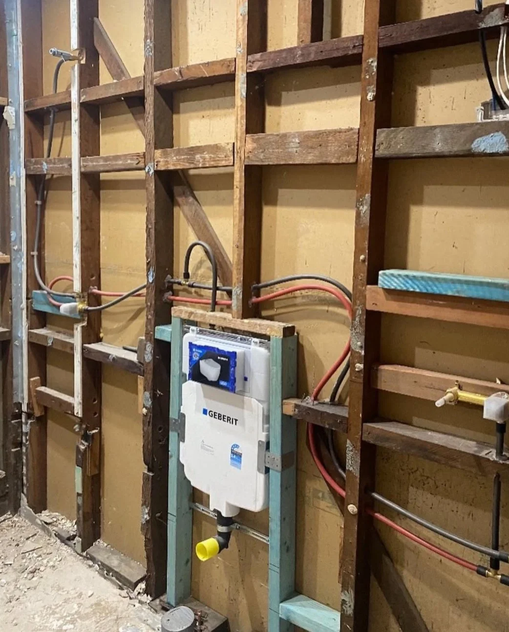Interior view of a construction wall with exposed wooden framing, plumbing pipes, and a Geberit in-wall toilet tank system being installed.